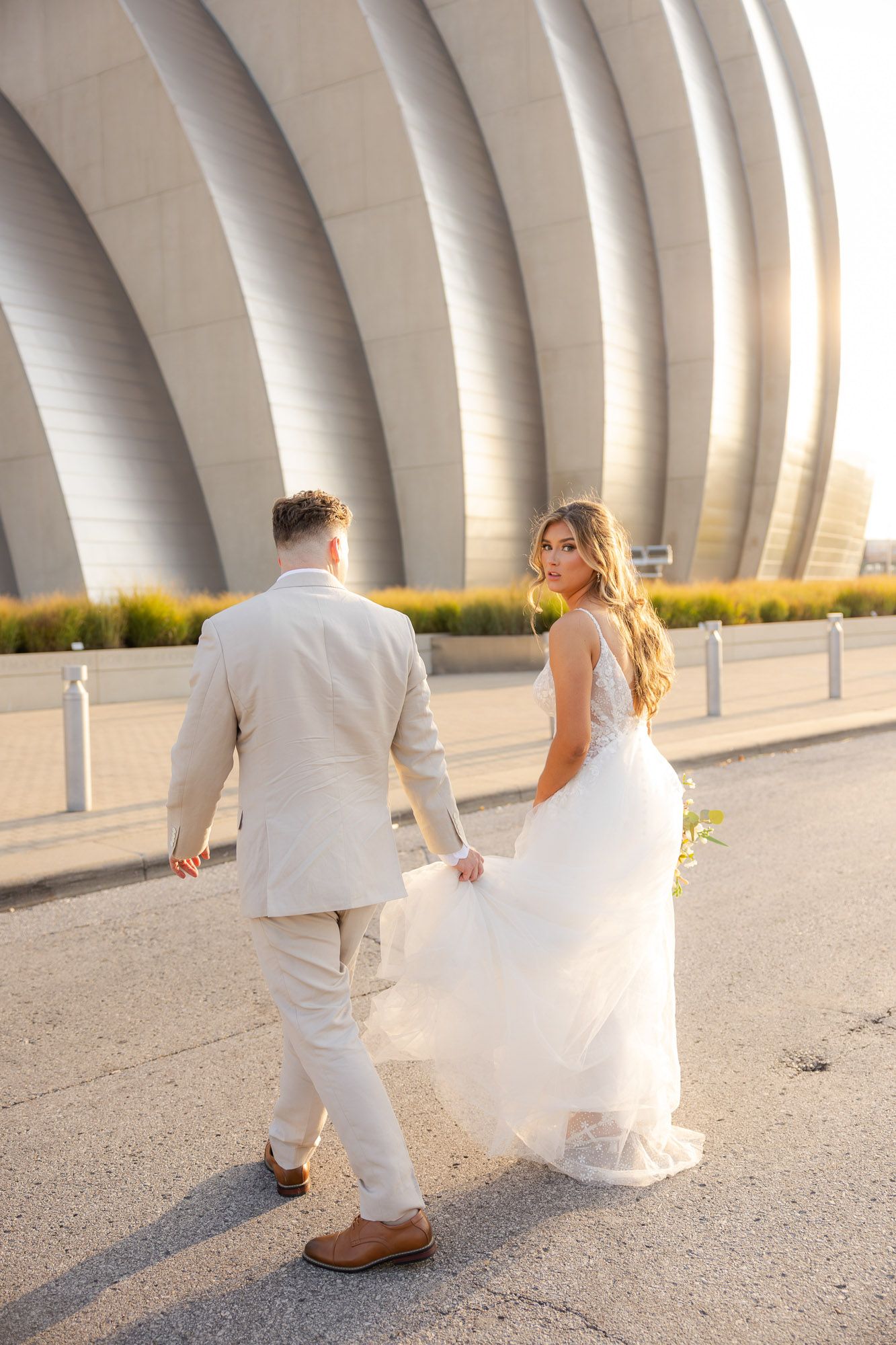 Bride in a flowing white gown and groom in a tan suit walking toward a modern architectural venue at sunset.