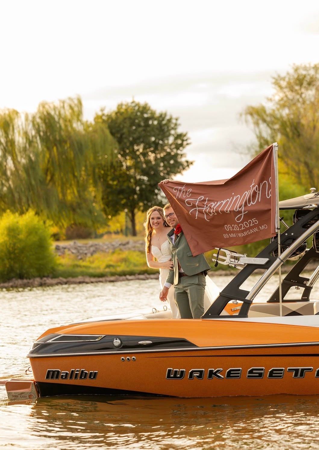 Couple posing on a Malibu Wakesetter boat on a lake, holding a custom flag at golden hour.