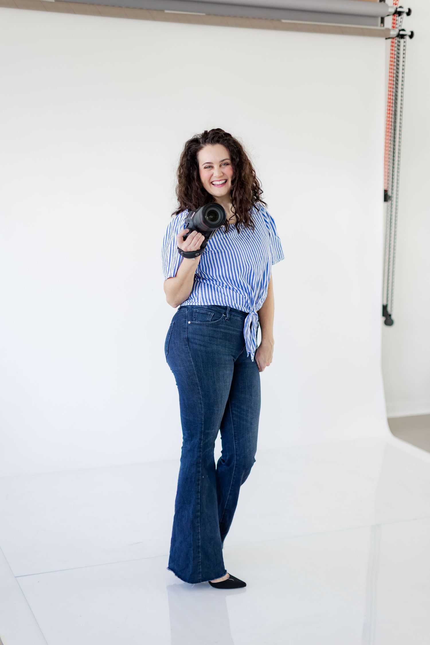 Happy curly-haired woman standing in photo studio holding camera wearing blue striped top and flare jeans.