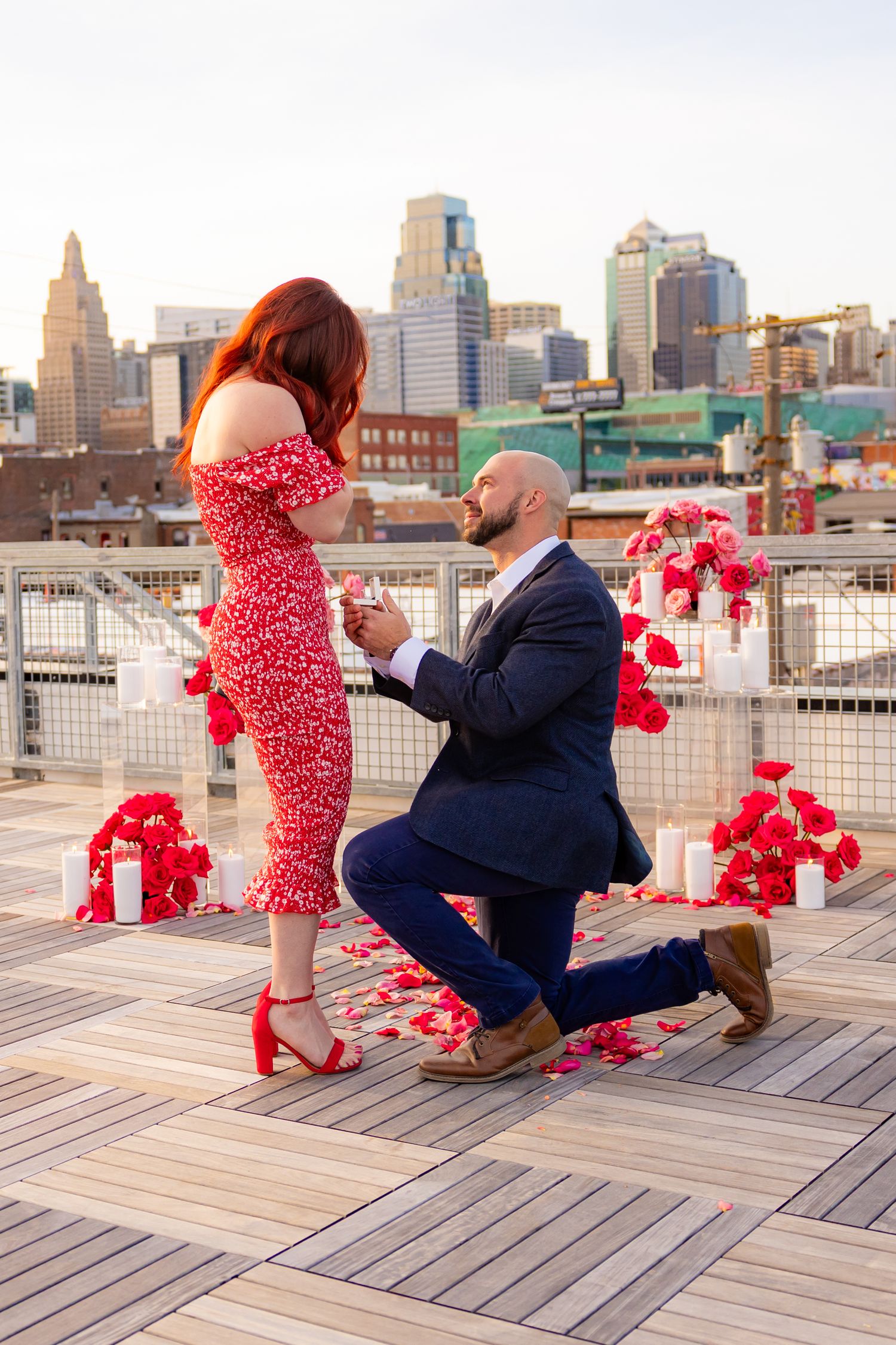 Man proposes on one knee to woman in red dress on rooftop with city skyline, rose petals and candles.