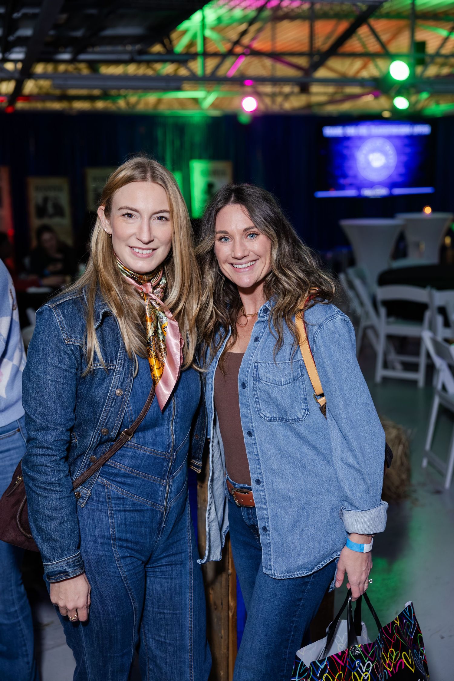 Two women in denim outfits smile together at a colorfully lit indoor event venue.
