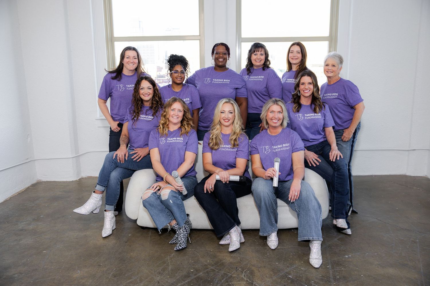 Group of eleven women in matching purple t-shirts posing together, some seated on a white couch, in a bright studio.