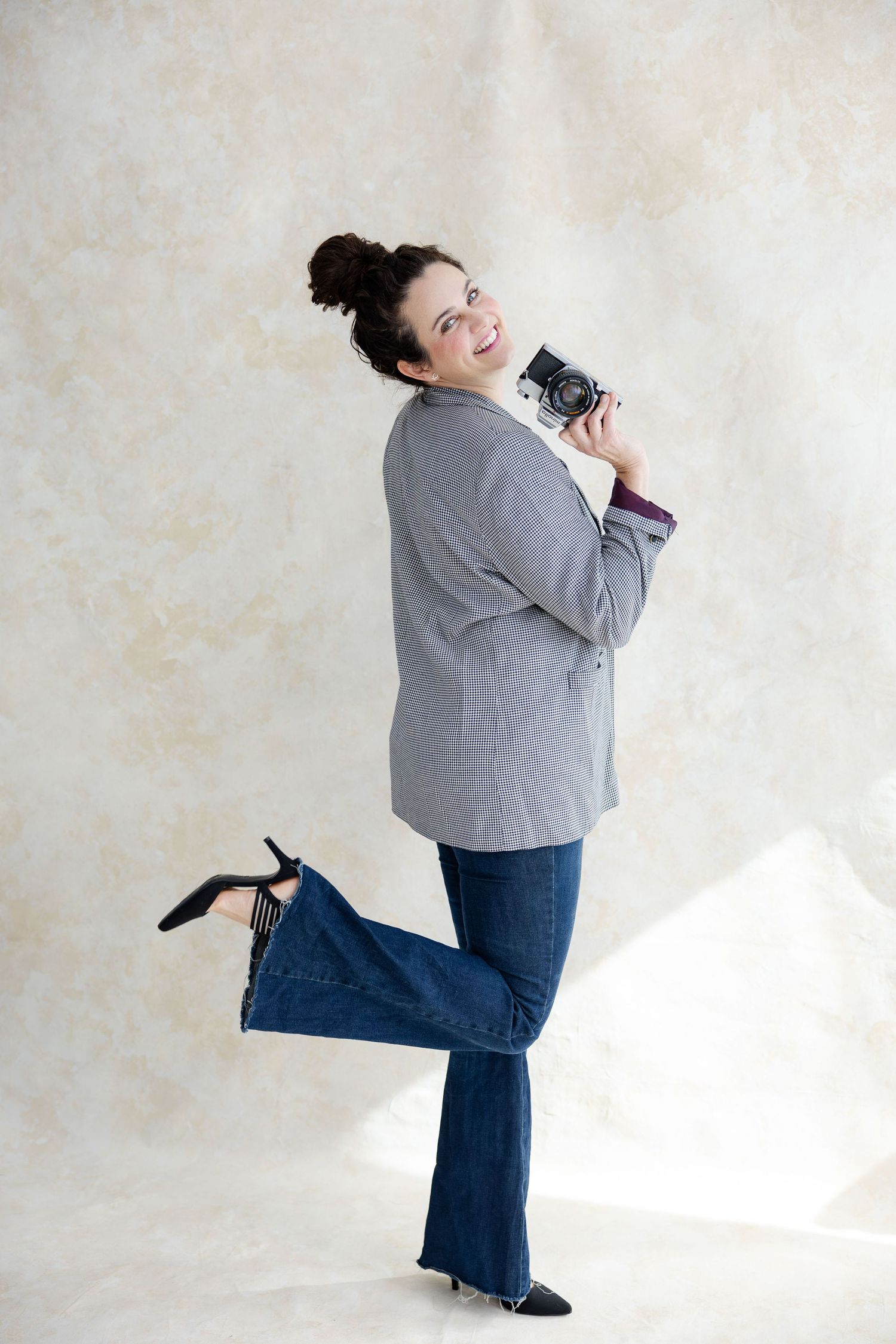 Joyful woman in gray blazer and jeans posing with vintage camera raised, one leg lifted playfully.