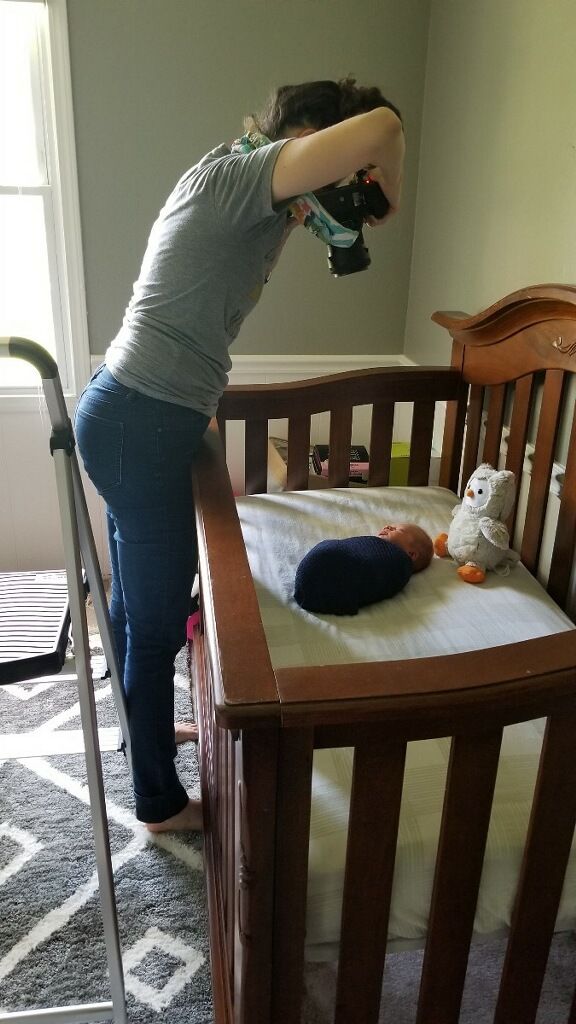Photographer leaning over wooden crib to photograph newborn baby lying on white mattress.