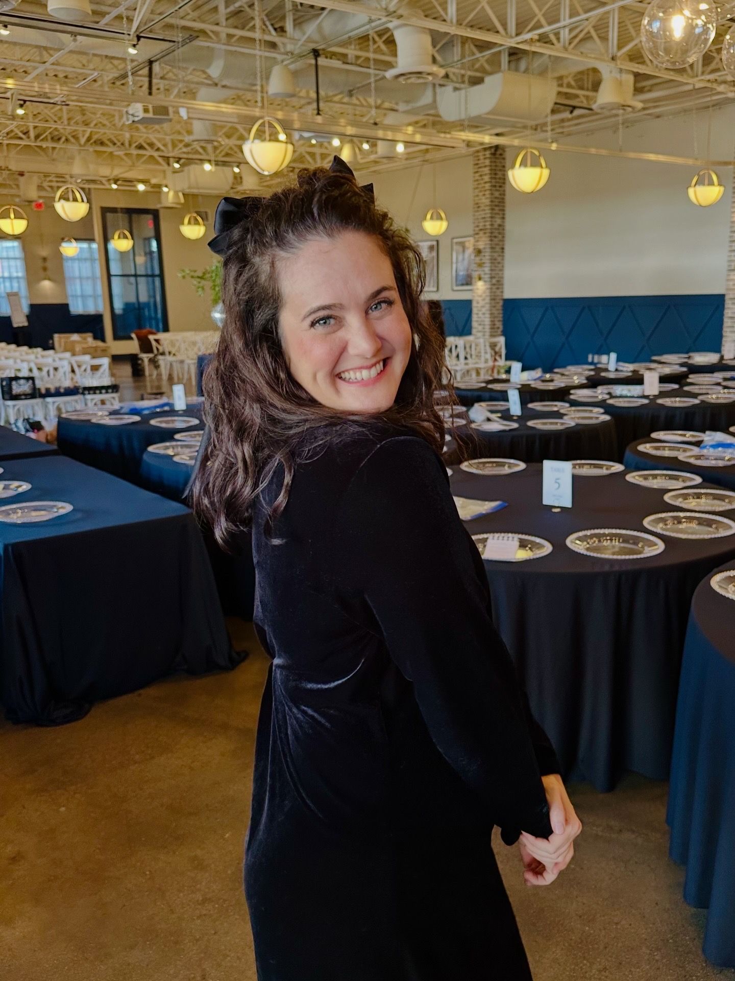 Smiling woman in black velvet dress turning back in a decorated banquet hall with round tables.
