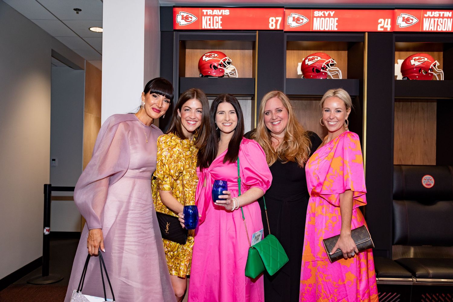 Five women in colorful formal attire posing in Kansas City Chiefs locker room.