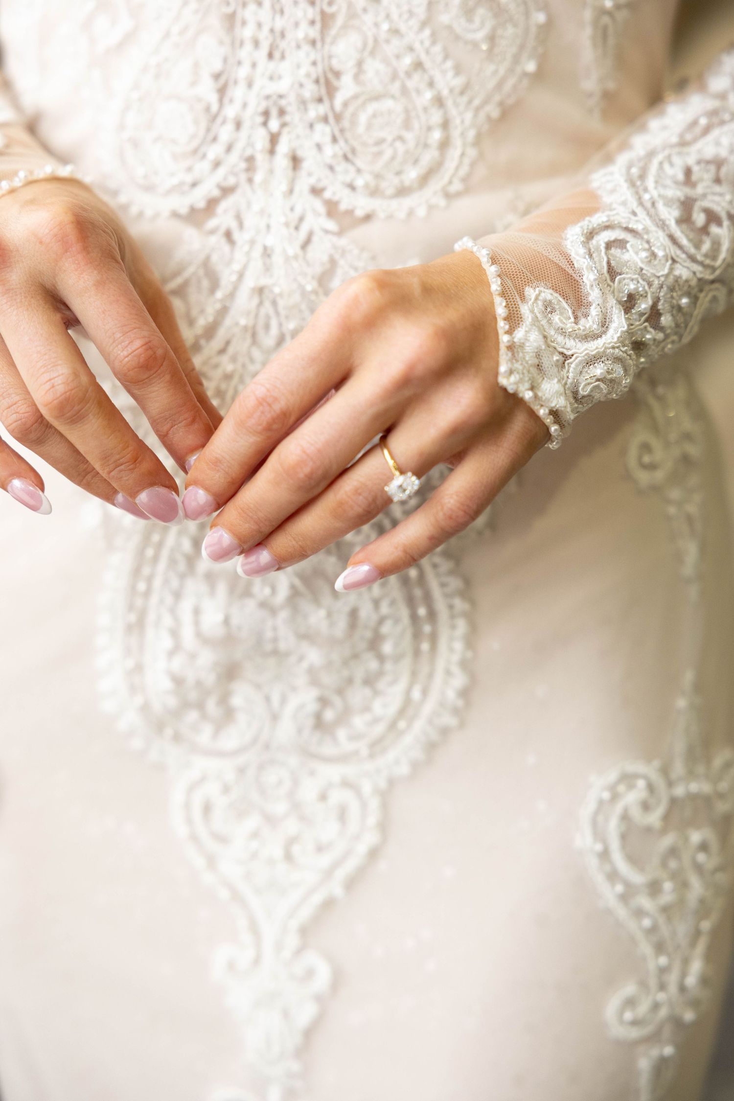 Close-up of a bride's hands wearing a diamond engagement ring on a lace wedding dress.