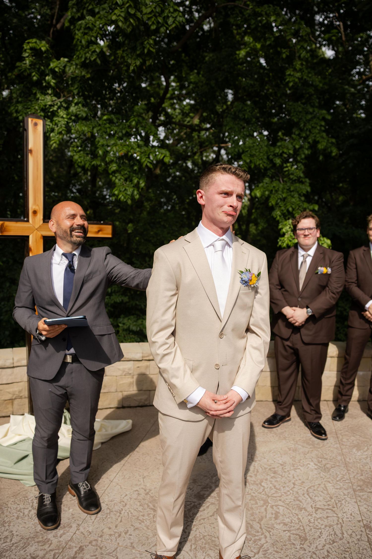 Groom in tan suit stands at outdoor wedding altar with officiant and groomsmen in brown suits beside wooden cross.