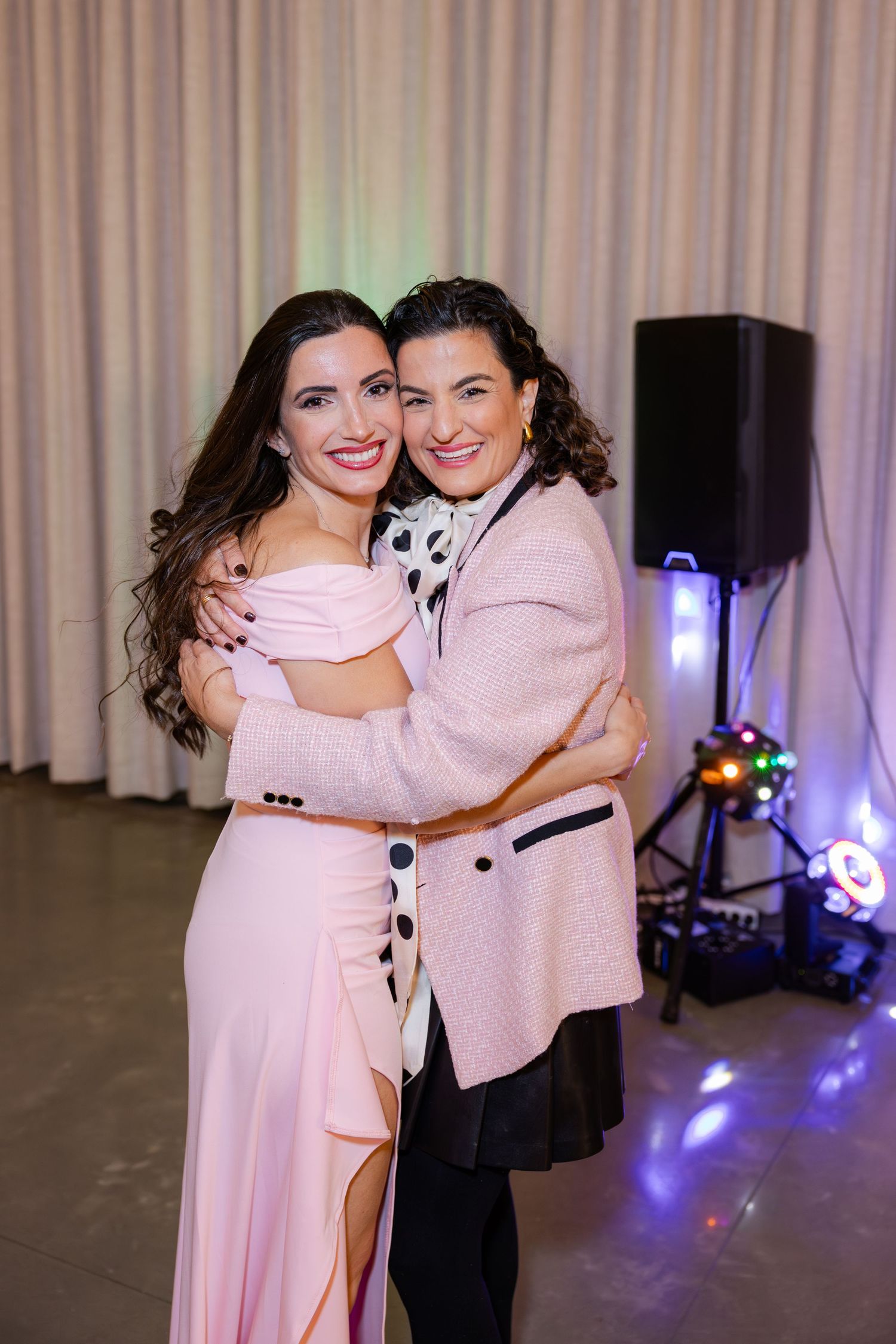 Two women in pink dresses embrace and smile warmly at a formal celebration event.