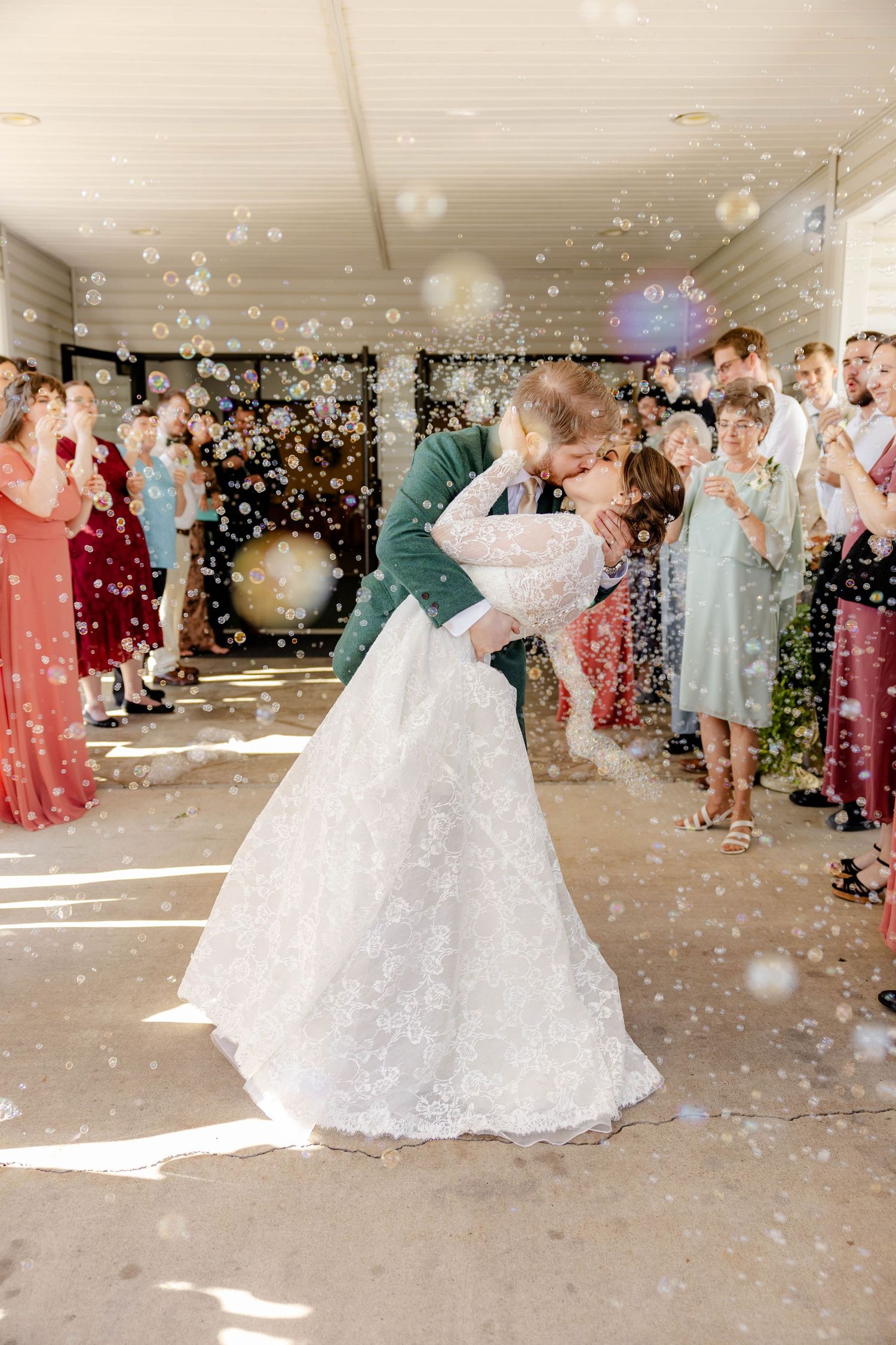 Groom dips bride for a kiss surrounded by bubbles and cheering wedding guests.