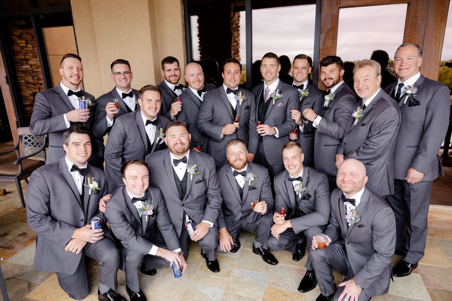 Large group of groomsmen in gray suits and bow ties pose together smiling at a wedding venue.