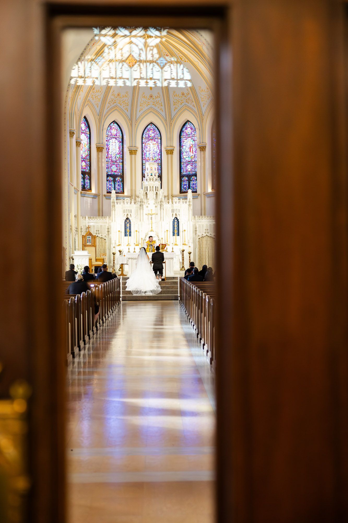 Wedding ceremony viewed through church doors with stunning stained glass windows and altar.