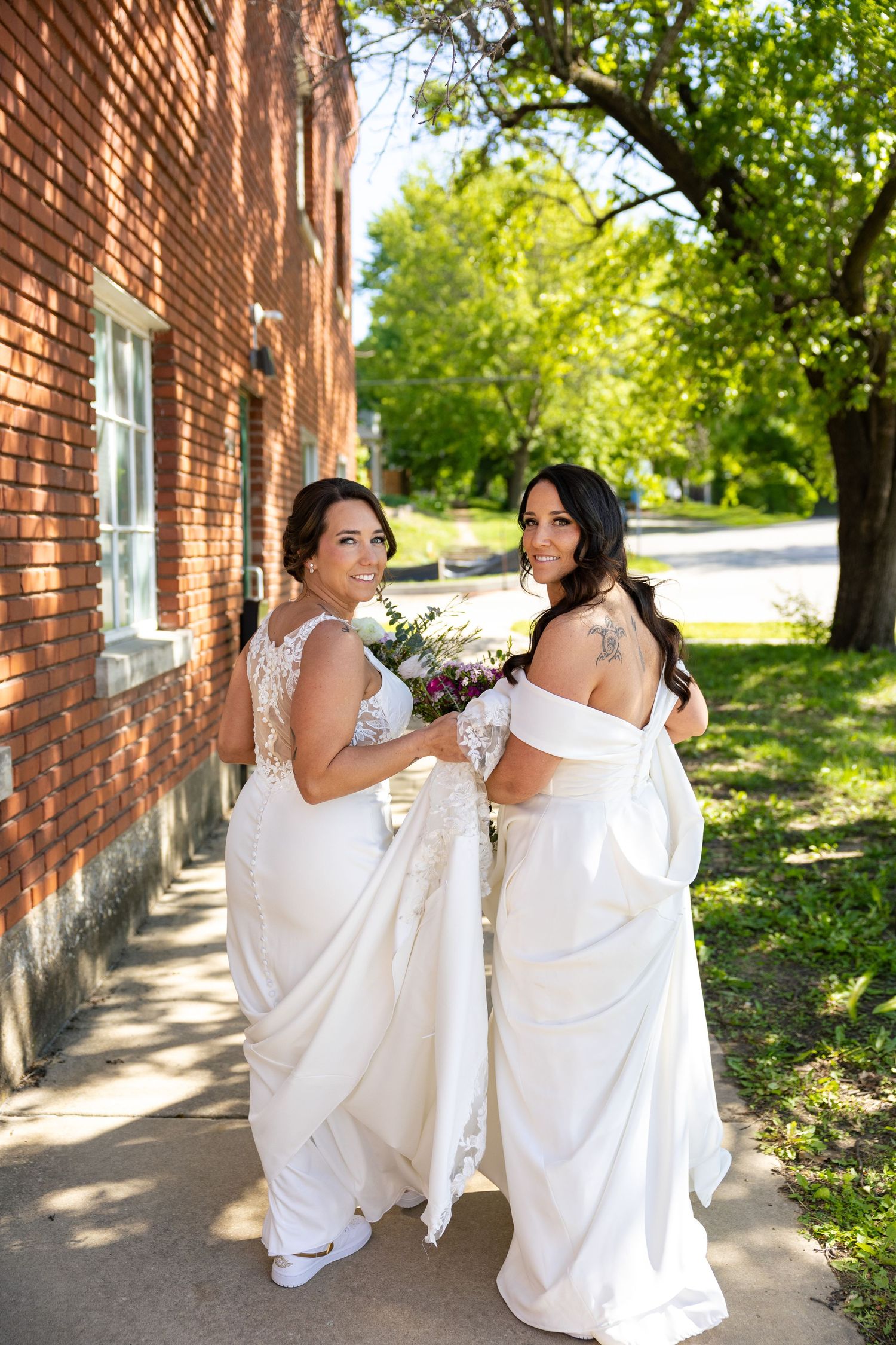 Two brides in white wedding gowns holding bouquets smiling outside brick building.