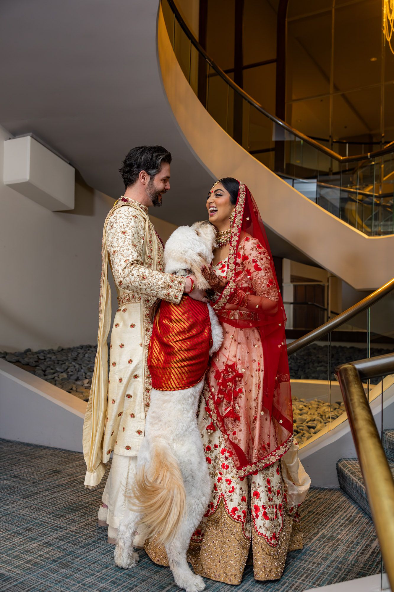 Indian wedding couple in traditional red and gold attire posing with white dog on staircase.
