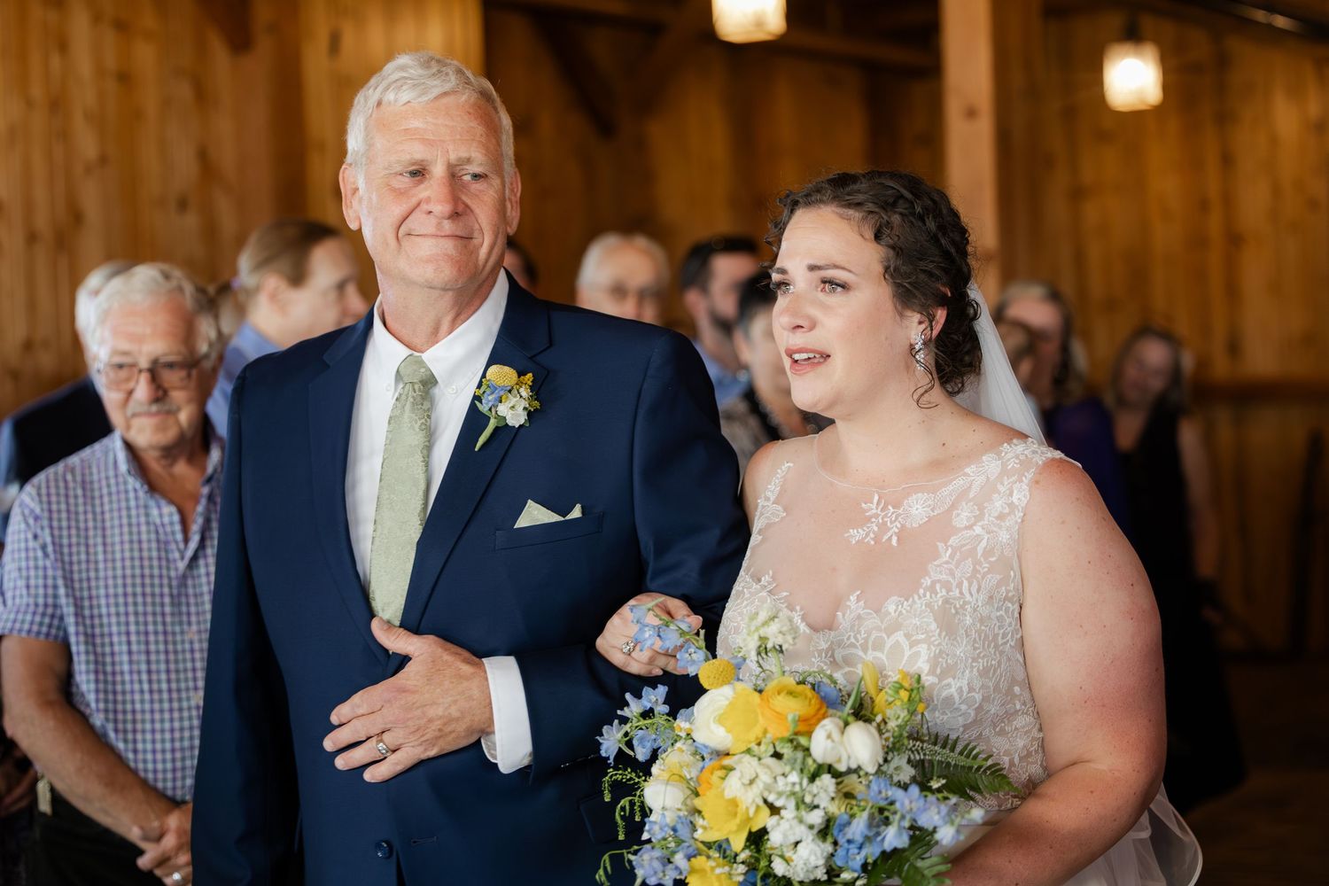 Bride holding yellow and blue bouquet walks down aisle with father in navy suit at rustic barn wedding ceremony.