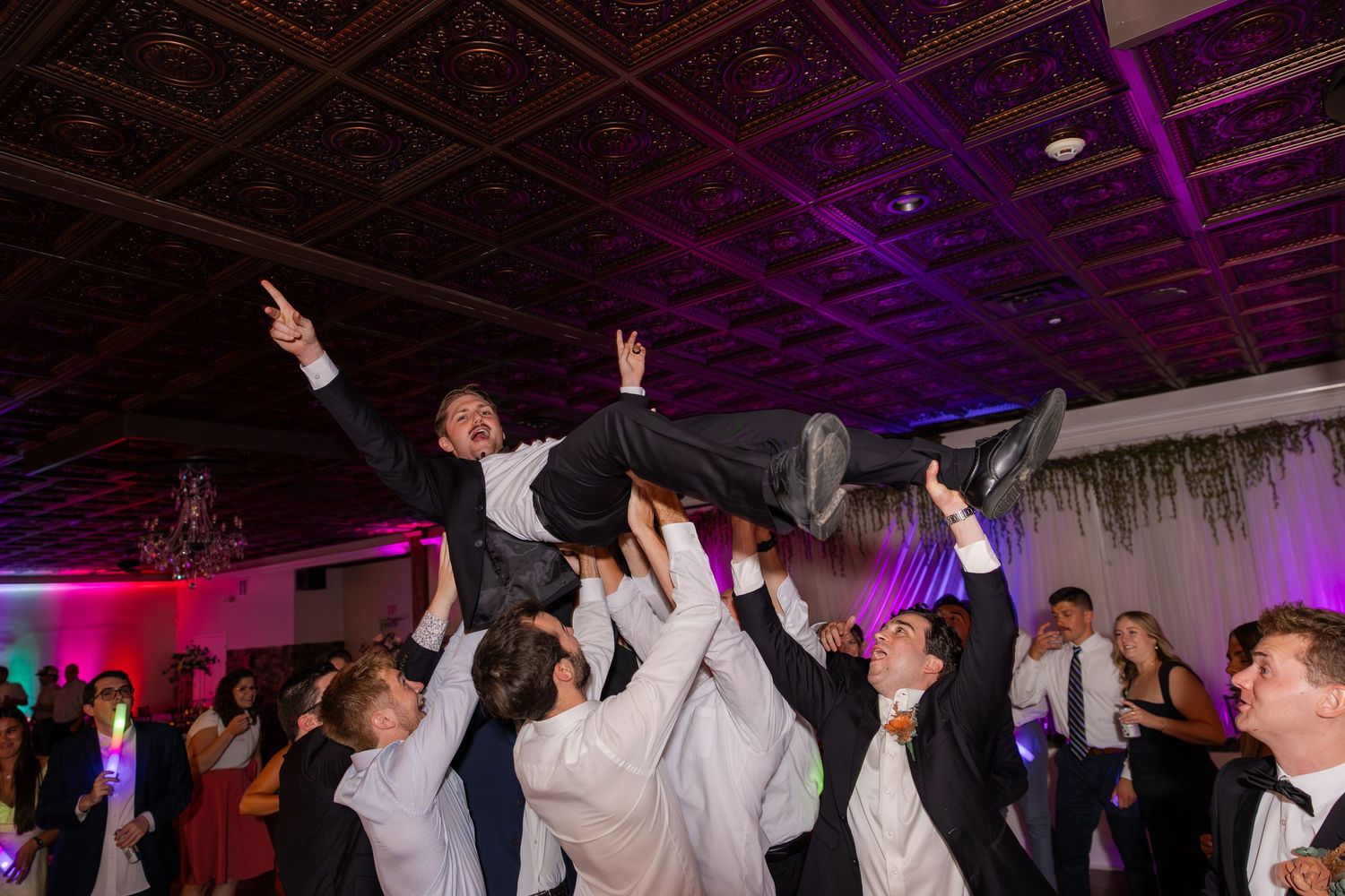 Groomsmen lift and carry groom above their heads during lively wedding reception dance party.