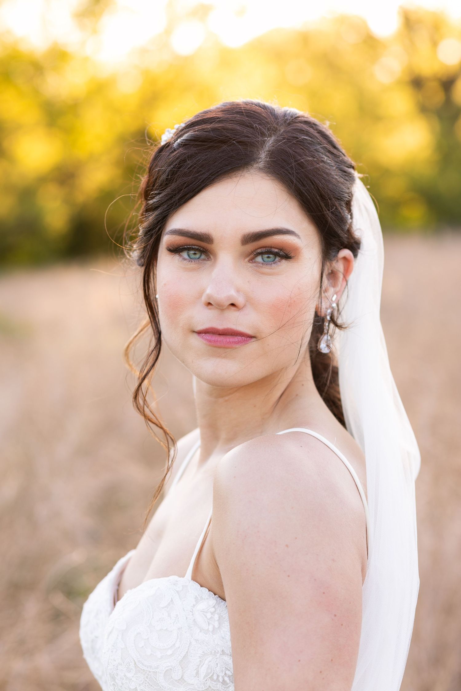 Beautiful bride with blue eyes and dark hair poses in a white lace gown with veil outdoors.