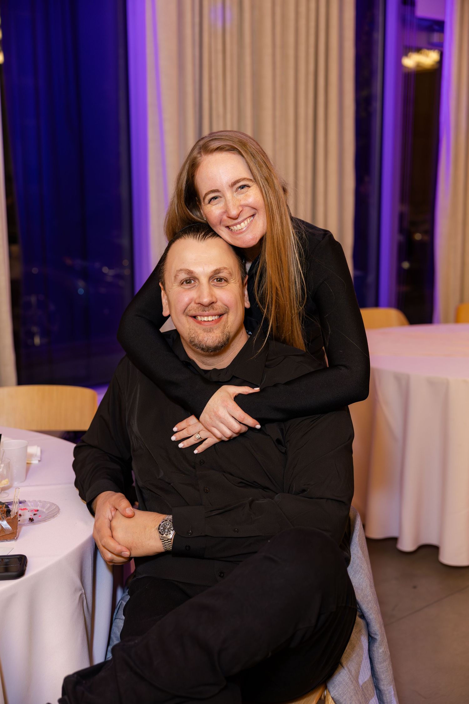 Smiling couple in black outfits posing together at a purple-lit evening event.