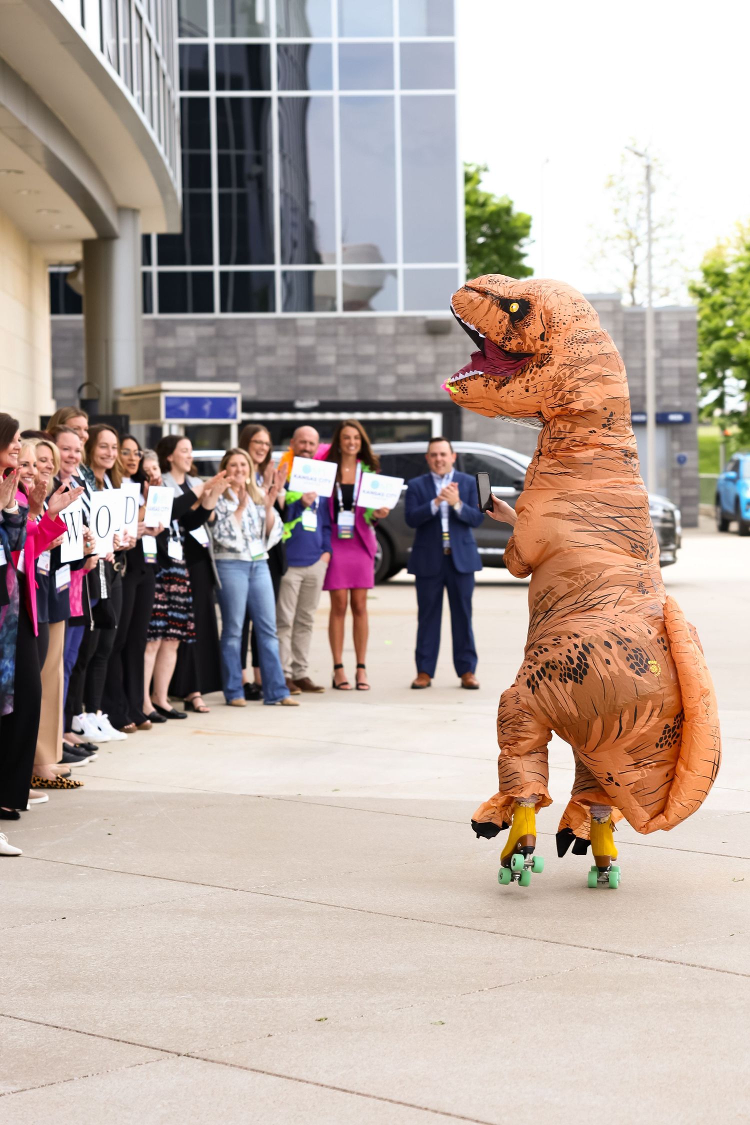 Person in inflatable T-Rex dinosaur costume on roller skates entertaining crowd outside modern office building.