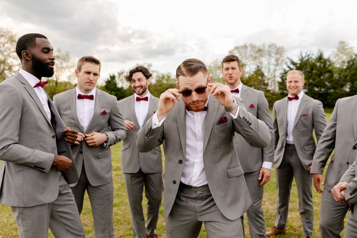 Groomsmen in gray suits with burgundy bow ties posing outdoors with groom adjusting sunglasses playfully.