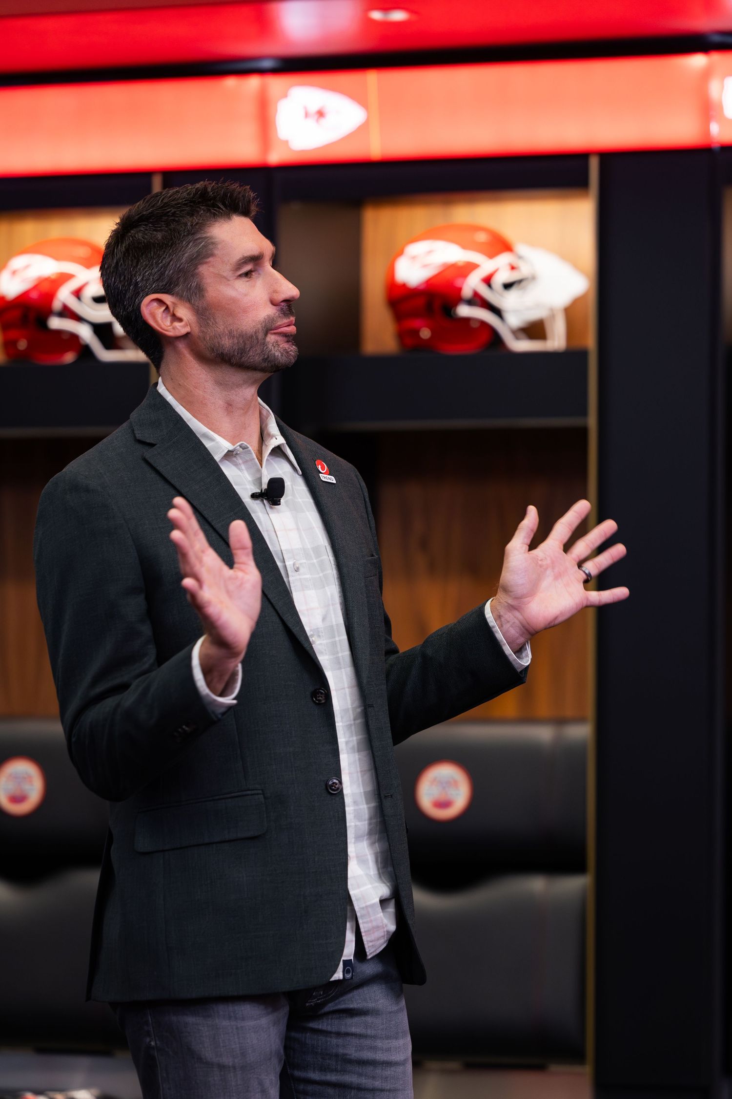 Man in blazer speaking on stage in a Kansas City Chiefs locker room setting.