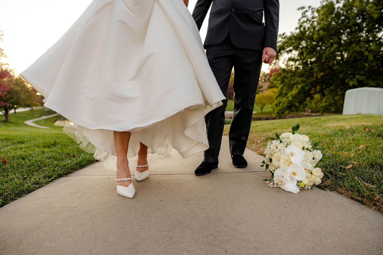Close-up of bride in white gown and groom in black suit walking on a pathway with white bouquet on ground.