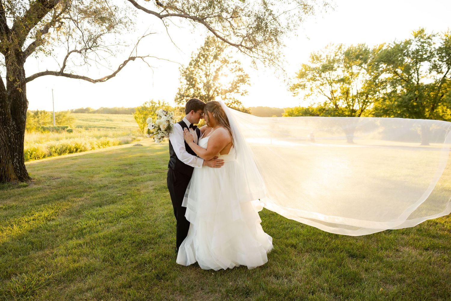 Bride and groom share a romantic moment at sunset, with her veil flowing in the wind on a lush green lawn.