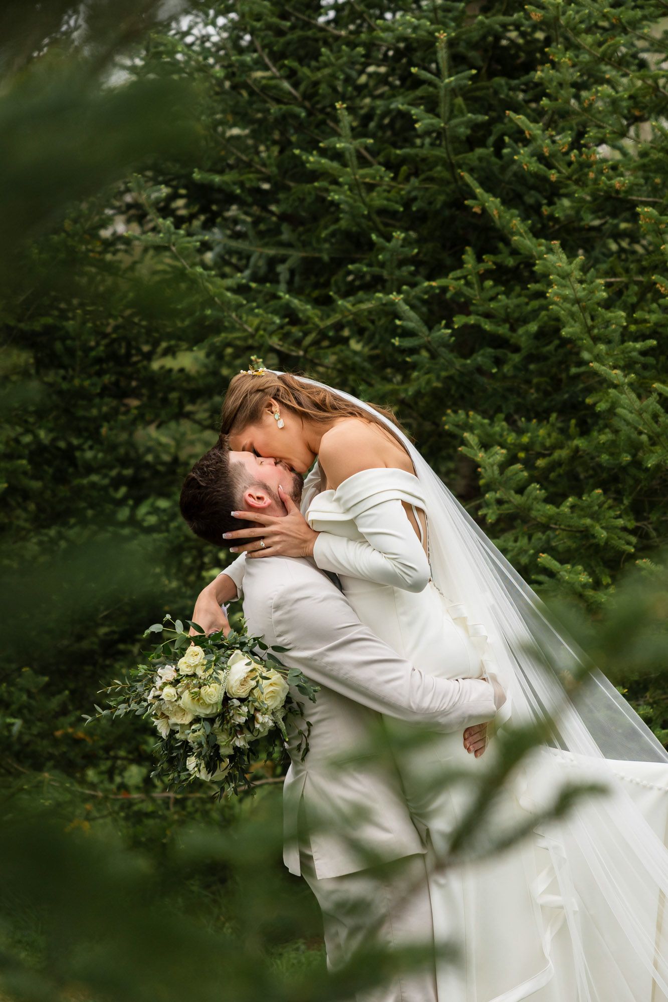 Bride and groom sharing a romantic dip kiss surrounded by lush green evergreen trees.