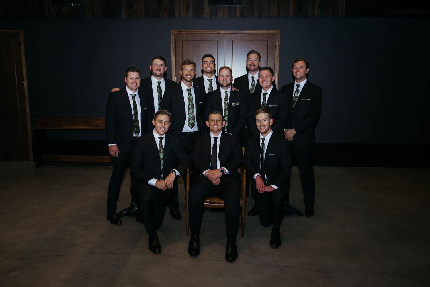 Groomsmen in black suits pose for a formal portrait, with groom seated in center in a dark room.