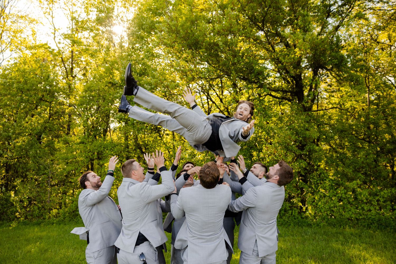 Groomsmen tossing groom into the air outdoors in gray suits with green trees in background.