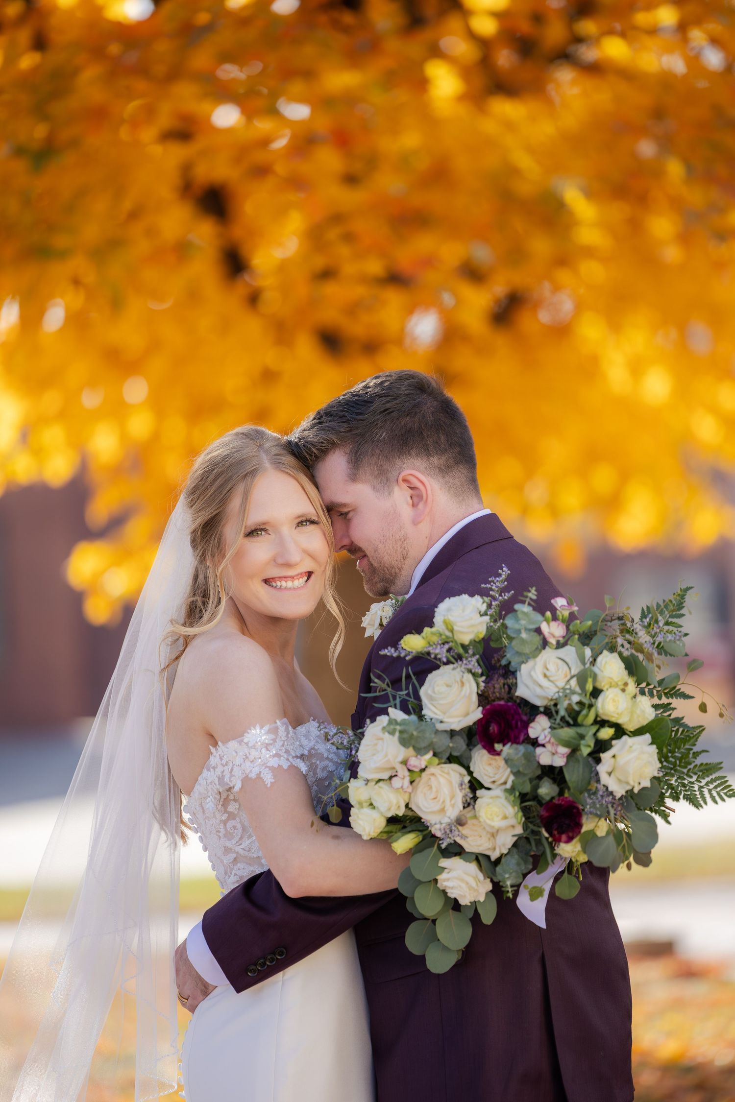 Bride and groom embrace under vibrant golden autumn foliage, bride holding lush white and burgundy bouquet.
