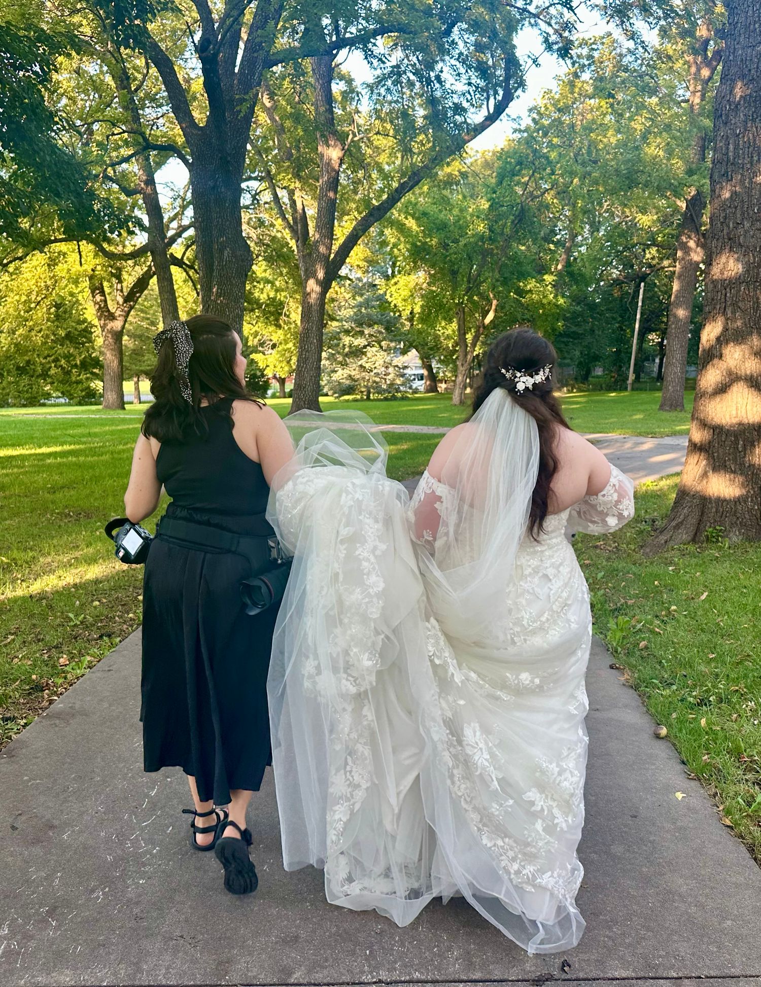 Bride in white lace gown and veil walks through a tree-lined park path with a companion in black.