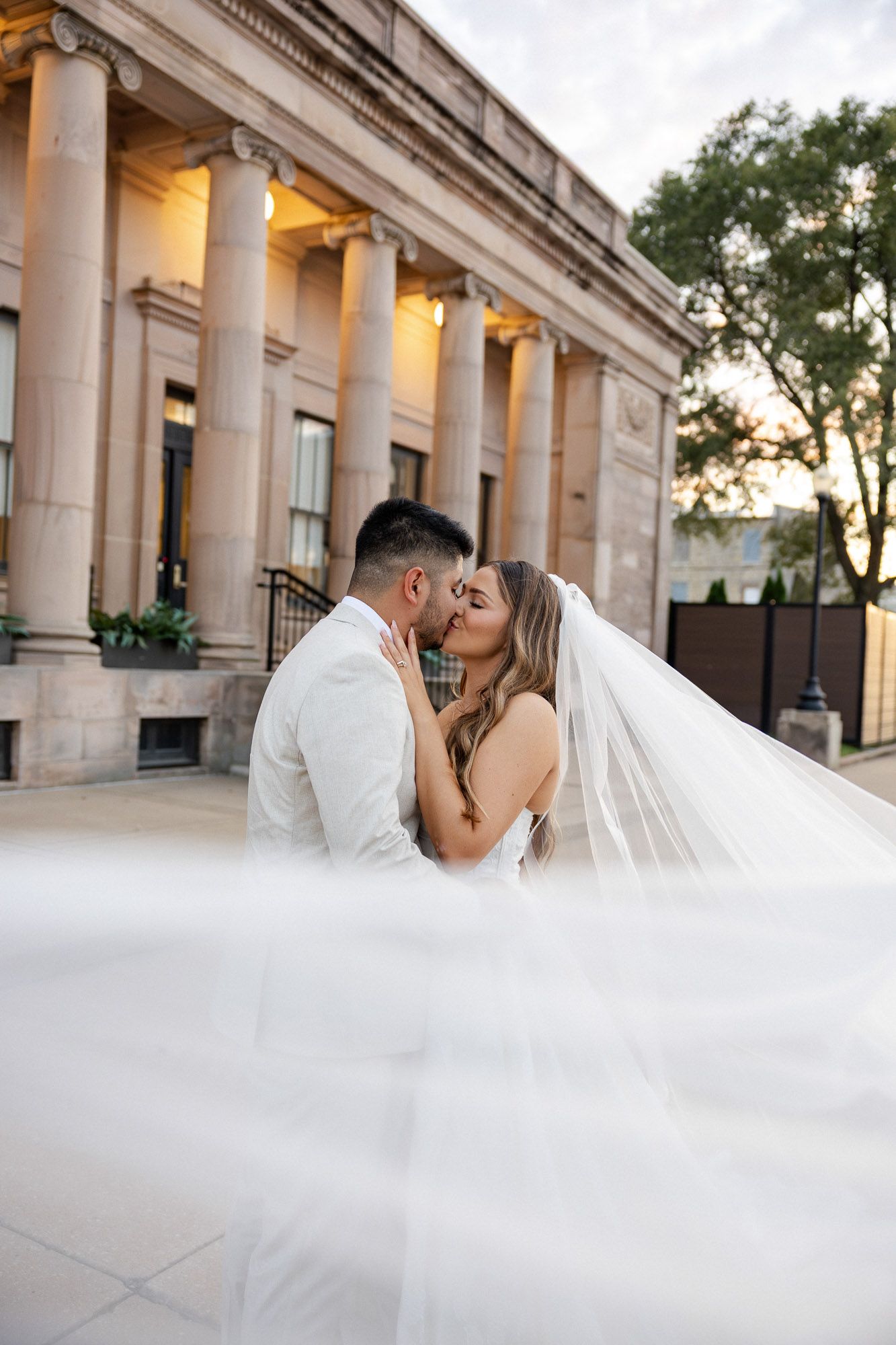 Bride and groom sharing a romantic kiss under a flowing veil in front of a grand columned building at sunset.
