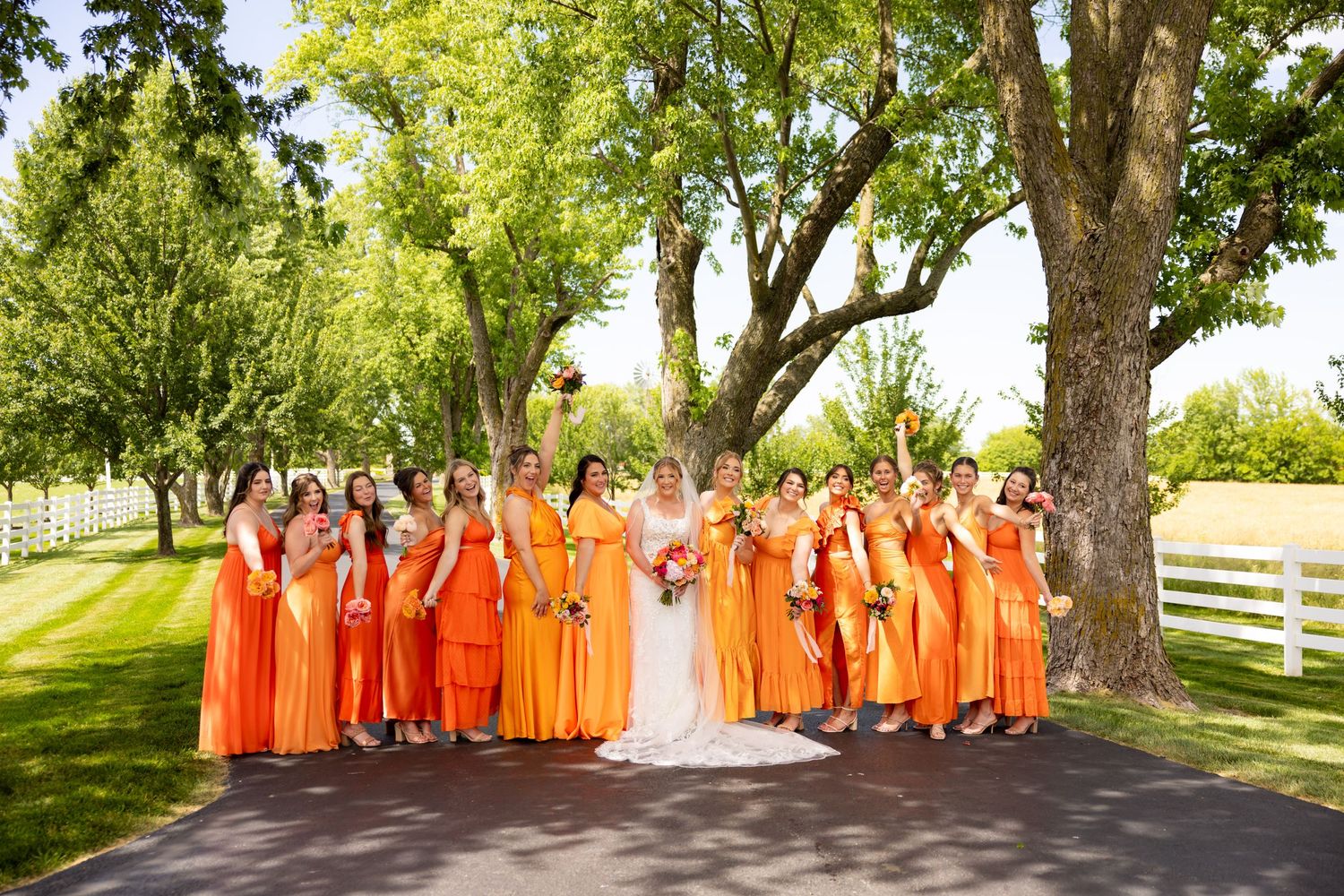 Large bridal party in orange dresses poses outdoors under green trees on a sunny wedding day.