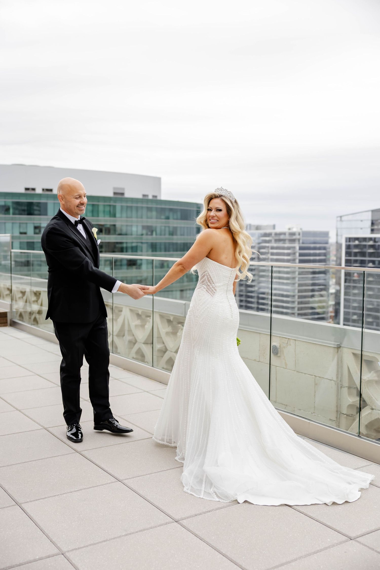 Bride in a fitted white gown with a tiara holding hands with a groom on a rooftop terrace.