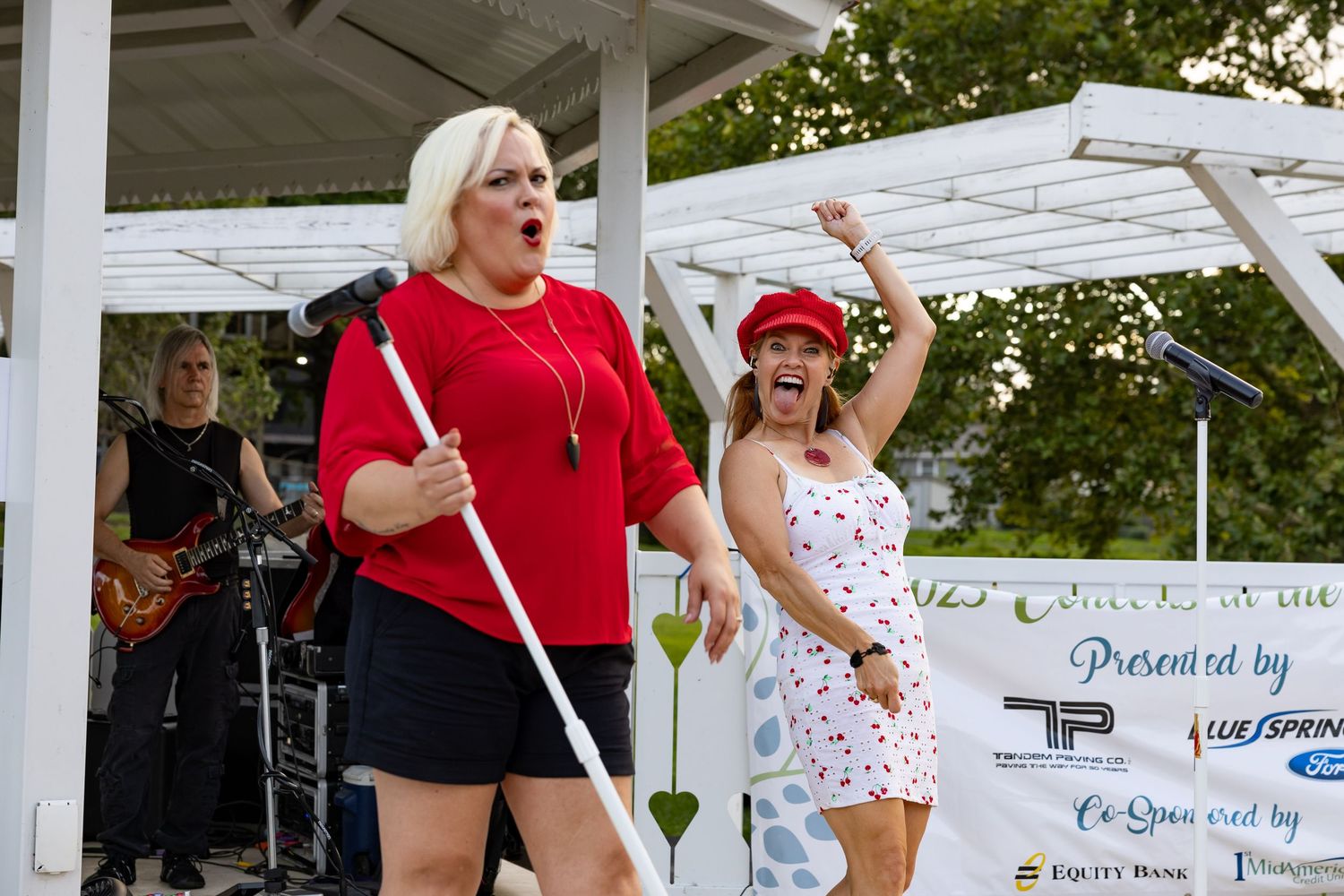 Two women performing energetically on outdoor stage, one holding a white cane.