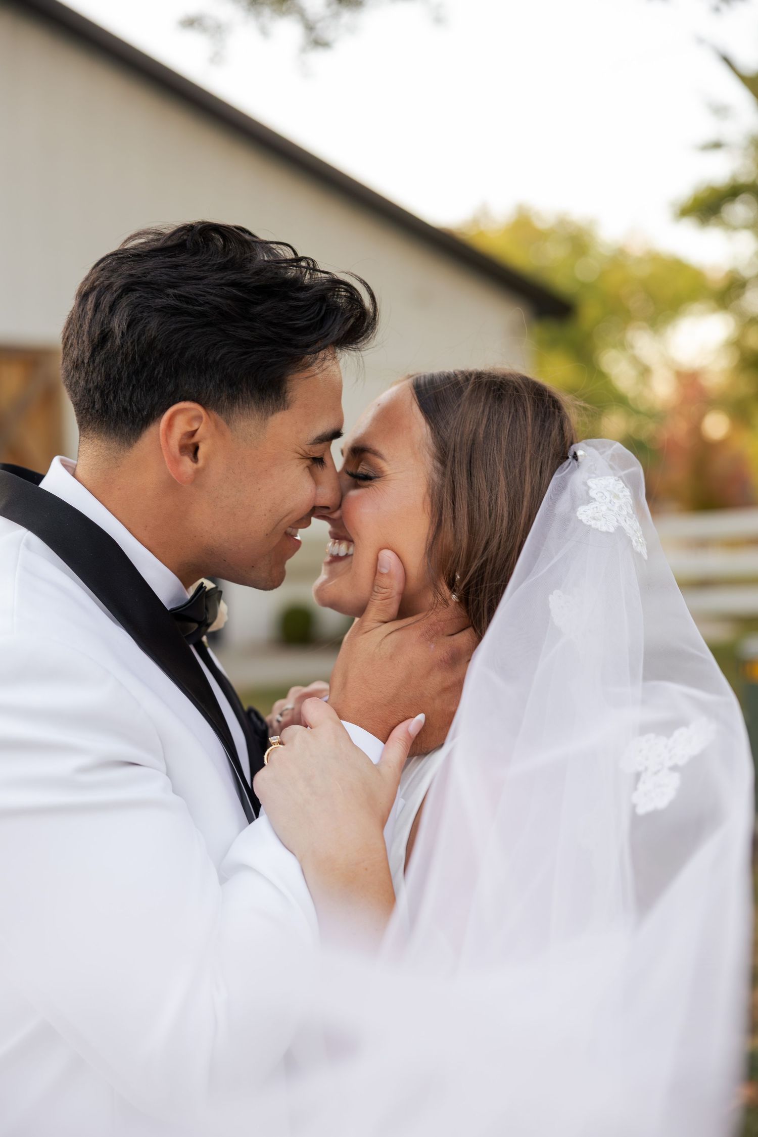 Groom tenderly holding bride's face as they lean in for a kiss outdoors at their wedding.