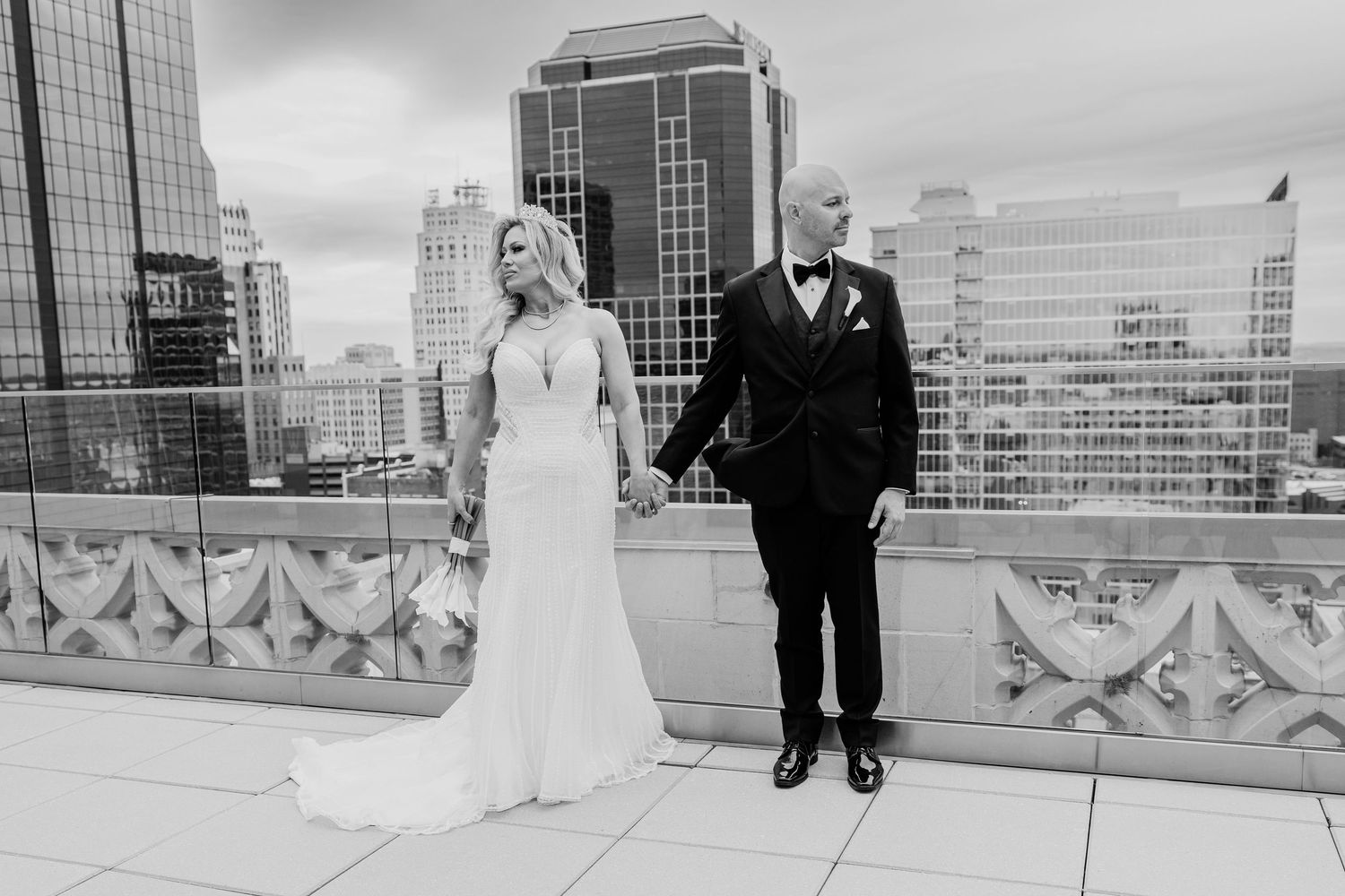Black and white photo of a bride and groom holding hands on a rooftop with city buildings behind.