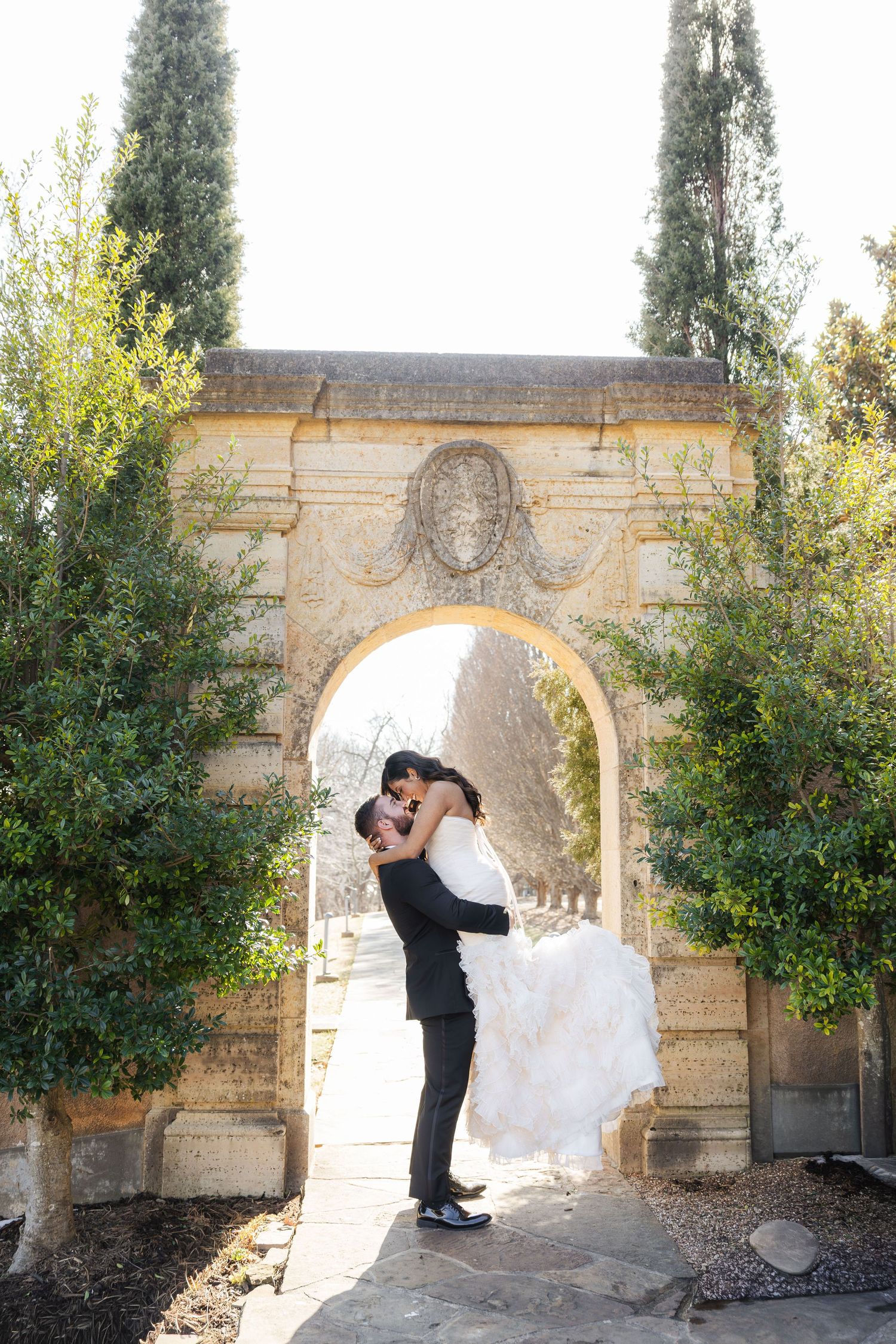 Groom lifts bride in wedding gown through ornate stone archway surrounded by tall cypress trees.