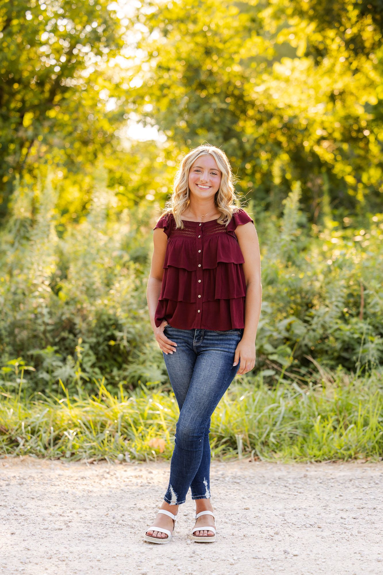 A smiling blonde teen girl poses in a maroon ruffled top and jeans on a sunlit gravel path.