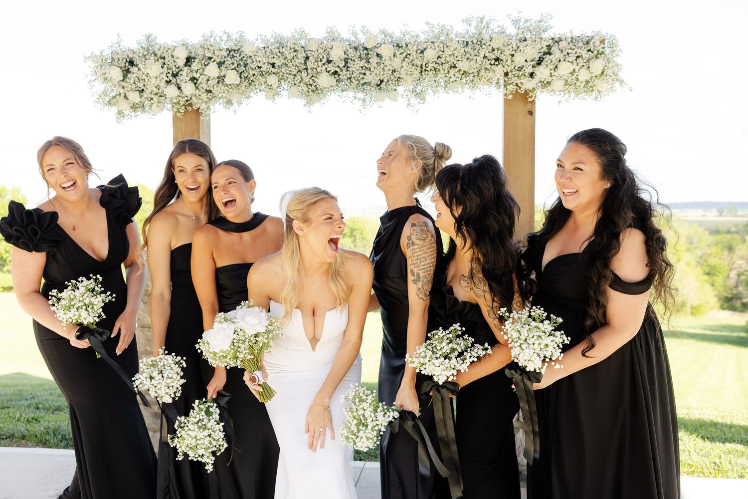 Bride with six bridesmaids in black dresses holding white bouquets under floral arch.