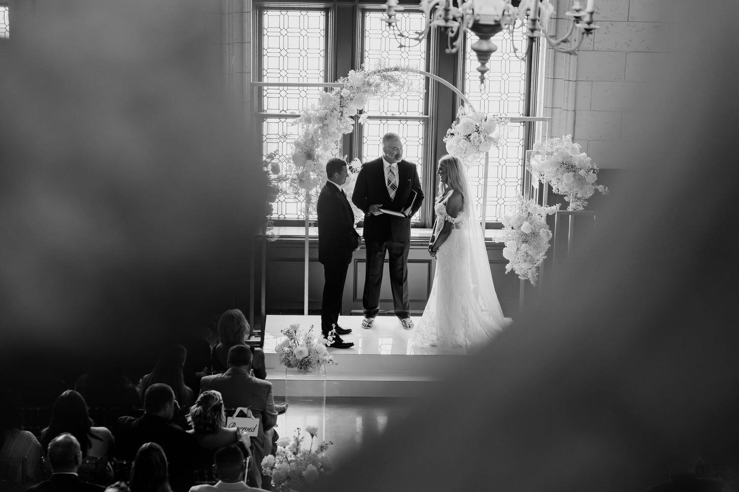 Black and white wedding ceremony with couple exchanging vows beneath floral arch in grand venue.