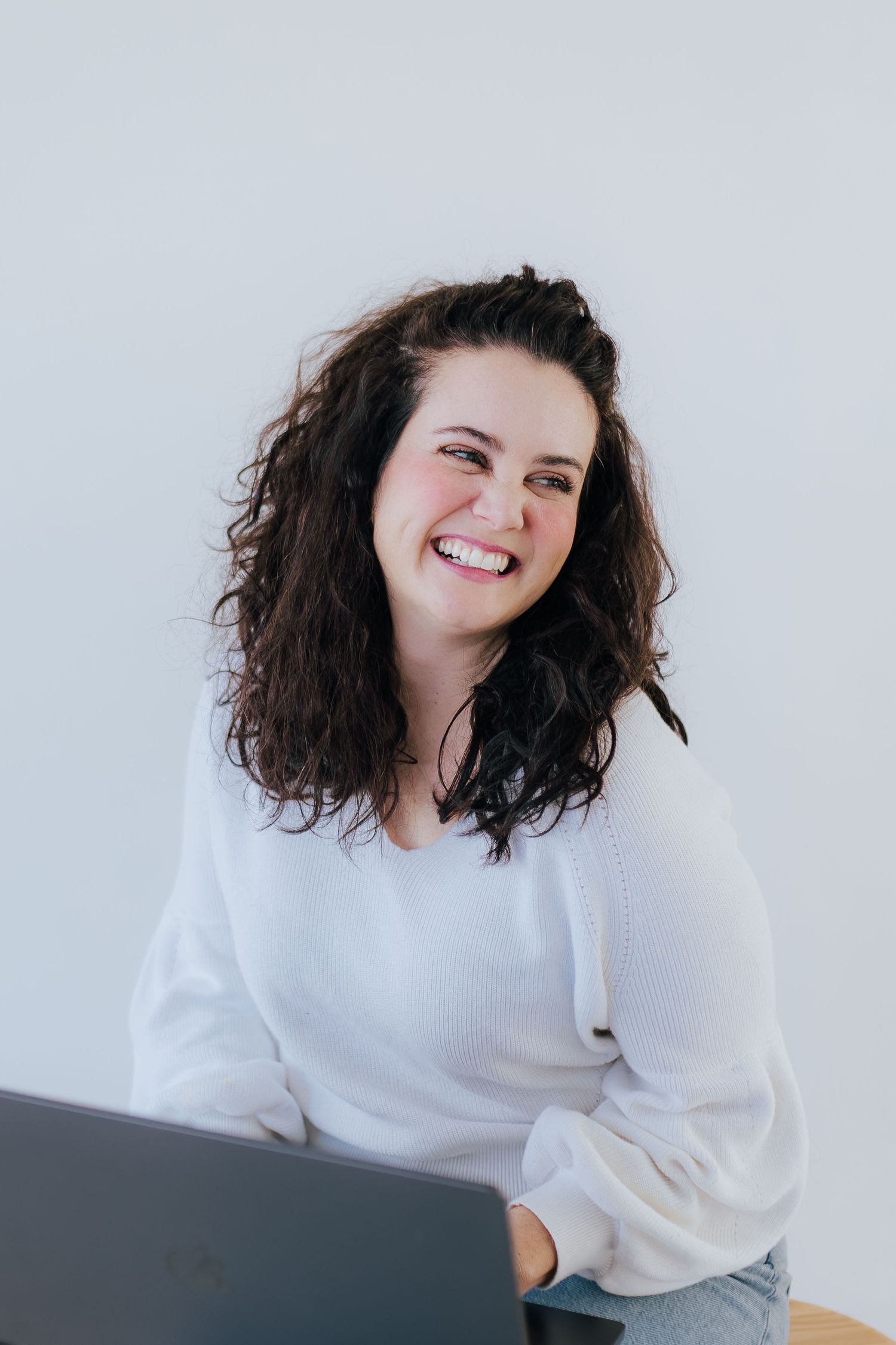 Smiling curly-haired woman in white sweater laughing while working on laptop against white background.