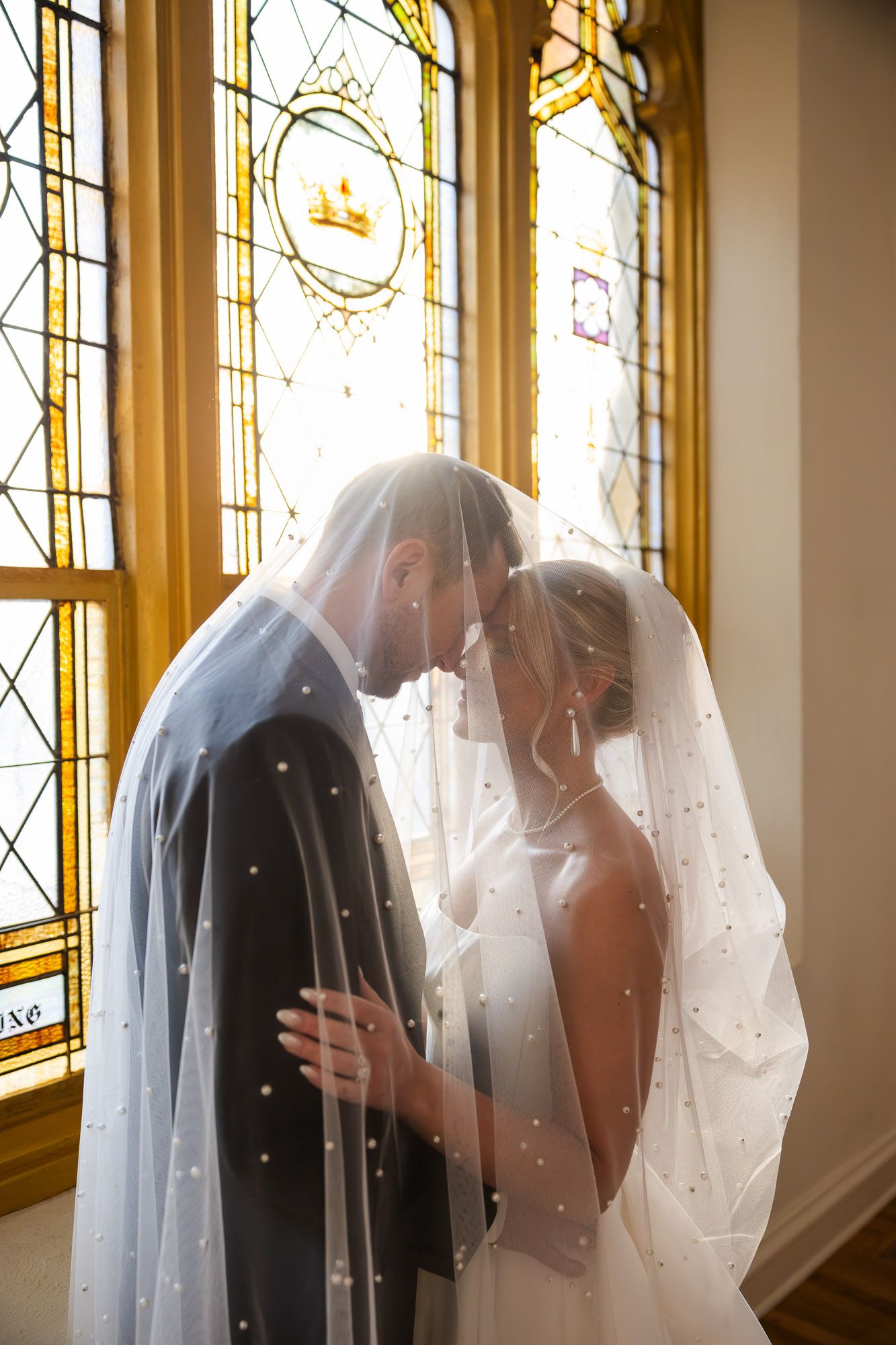 Bride and groom share intimate moment under pearl-adorned veil by stained glass window.