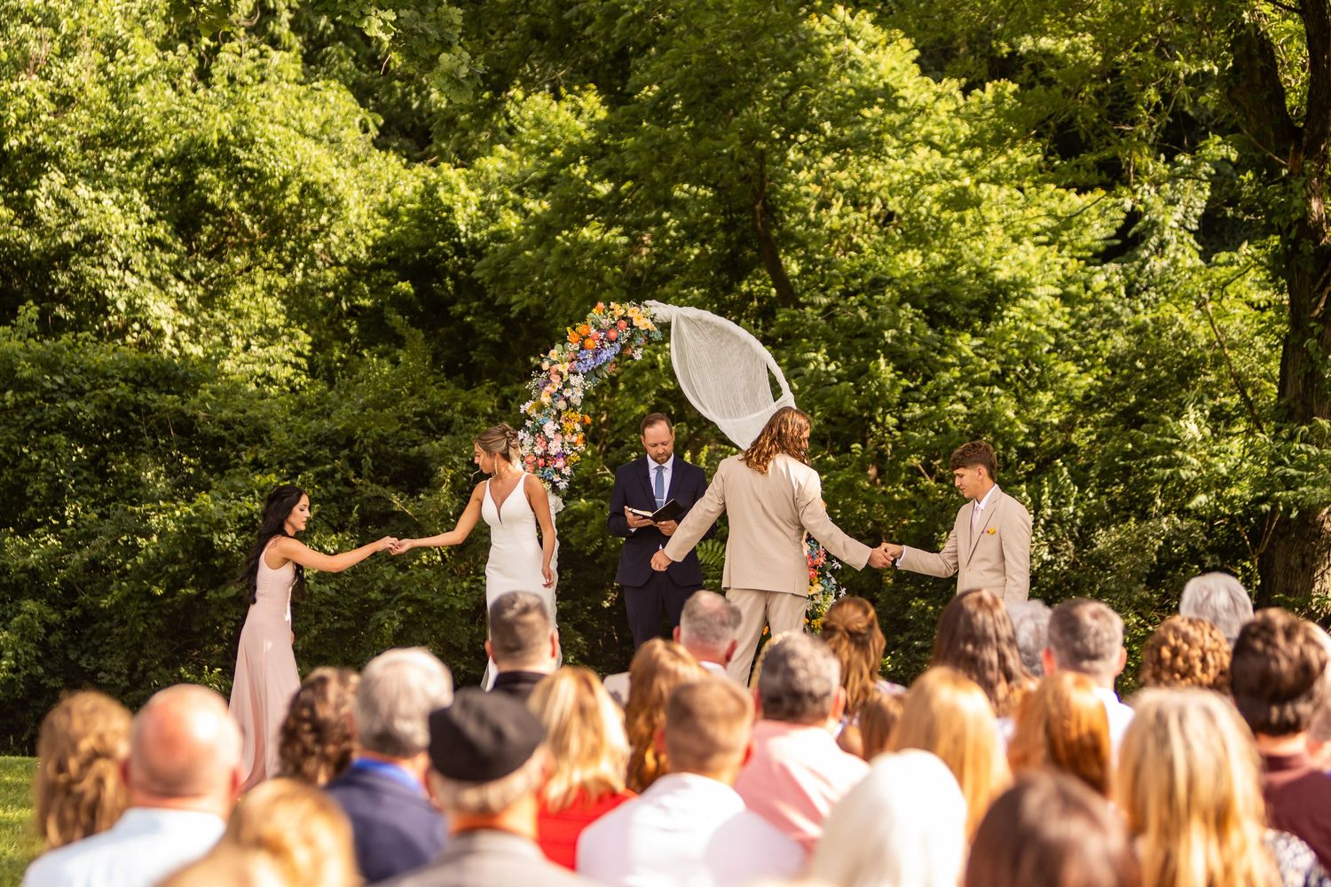 Outdoor wedding ceremony under floral arch with bridal party holding hands in lush green garden.