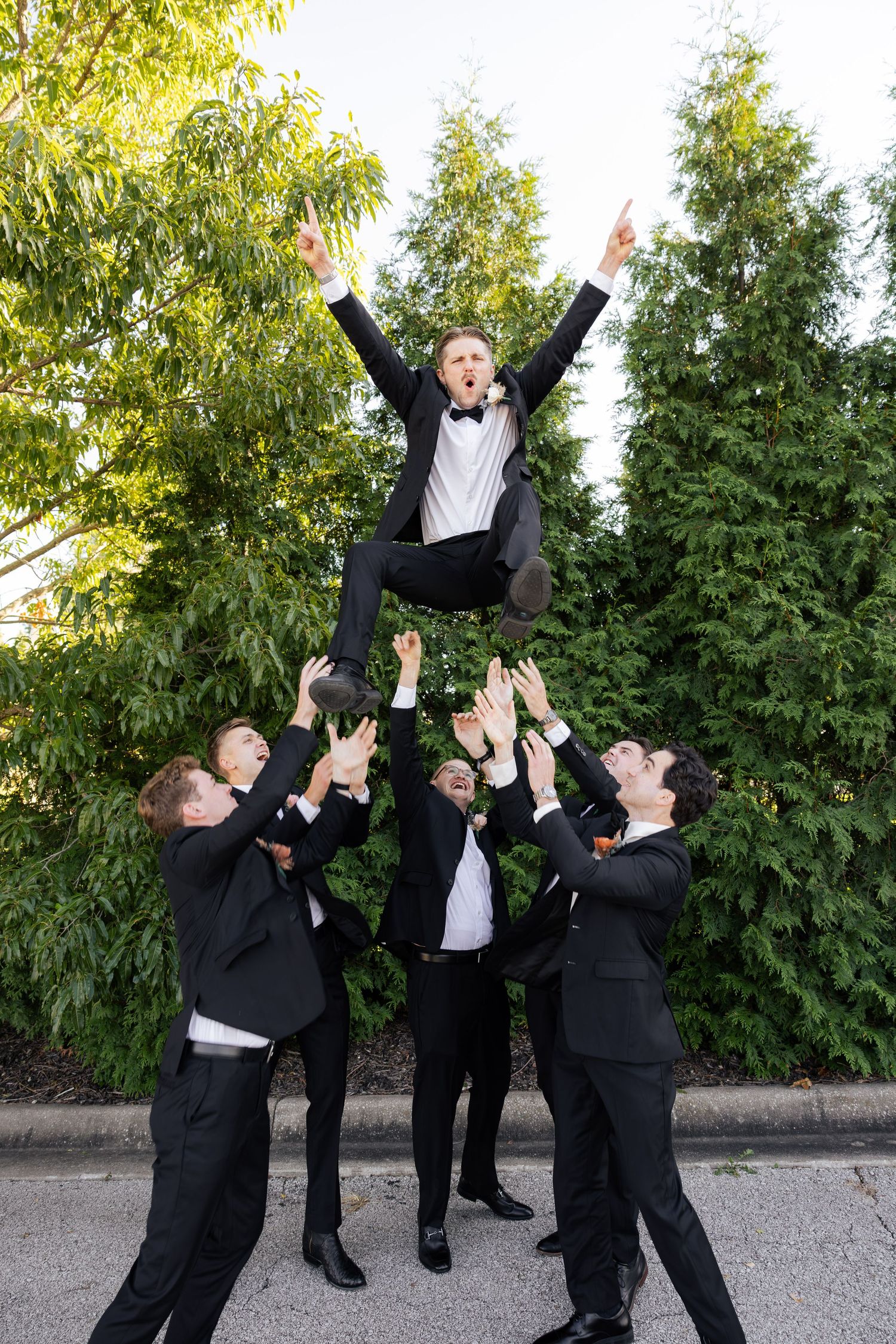 Excited groom is lifted into the air by cheering groomsmen outside wedding venue.