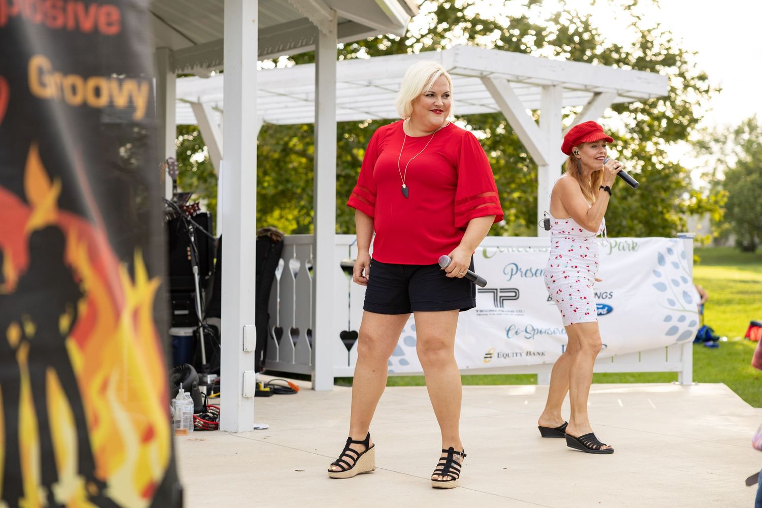 Two women performing on an outdoor stage, one in red top and black shorts.