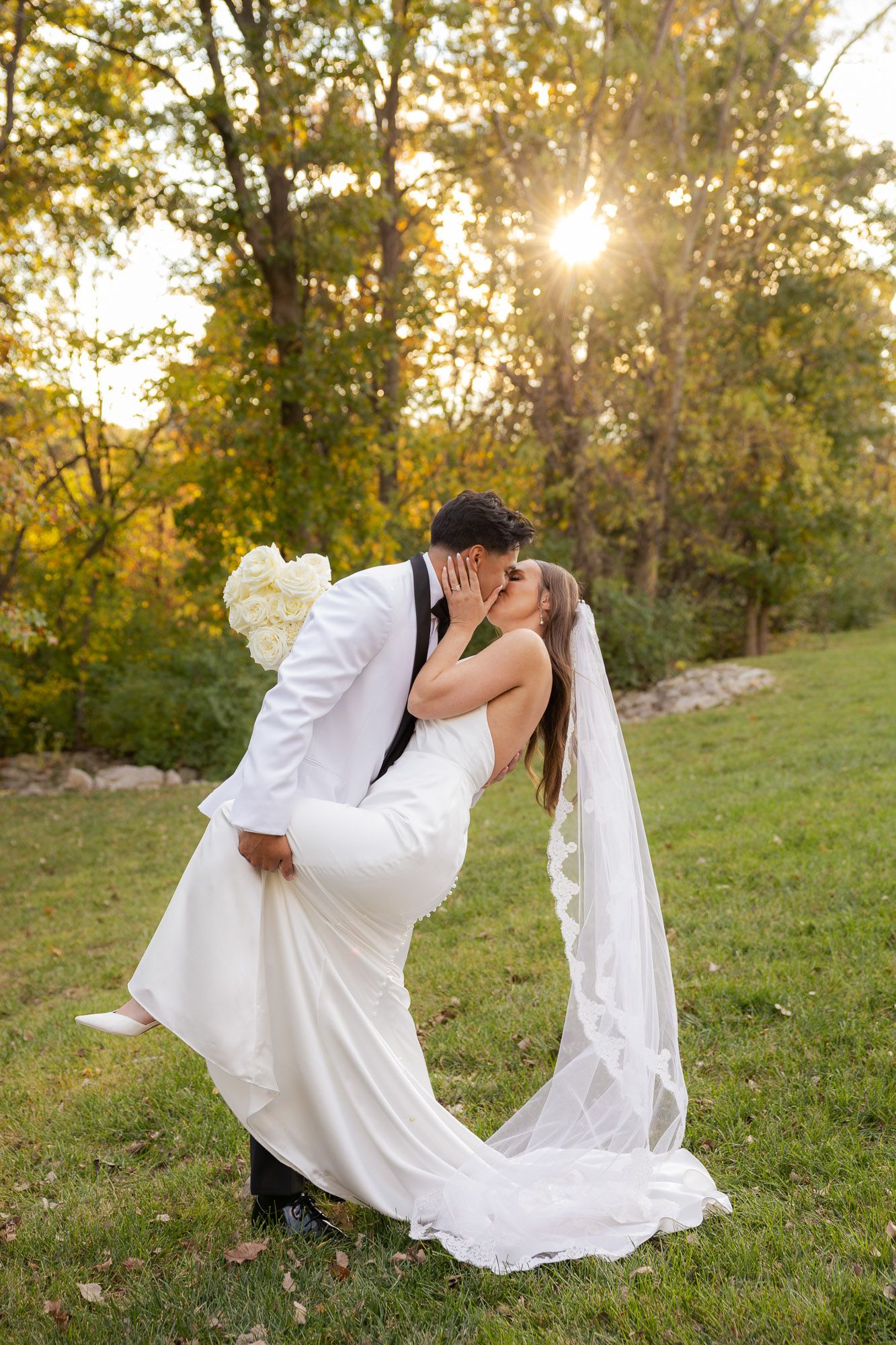 Bride and groom share a romantic dip kiss at golden hour in a lush green outdoor setting.