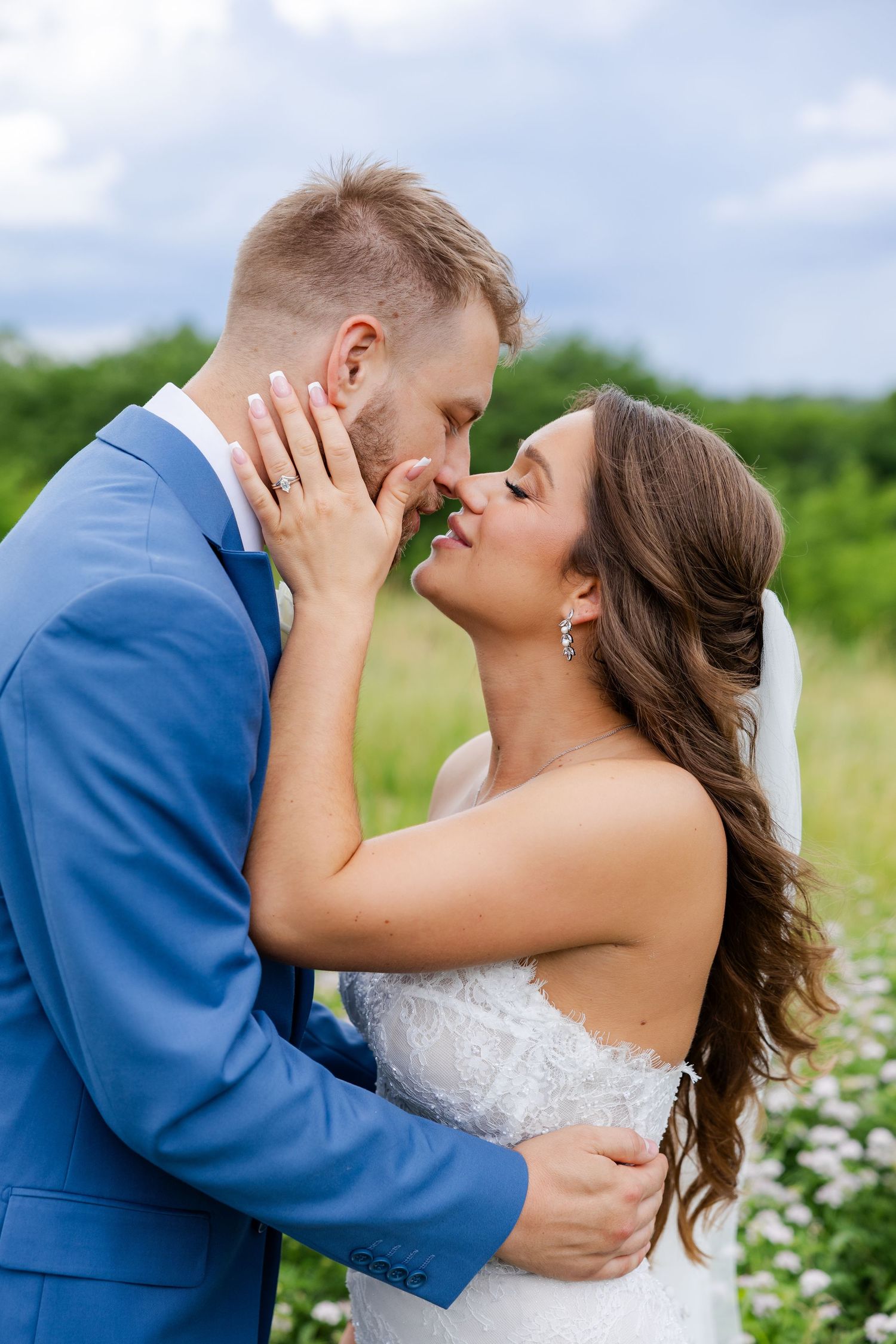 A bride and groom share a romantic almost-kiss outdoors, dressed in a blue suit and white lace wedding gown.