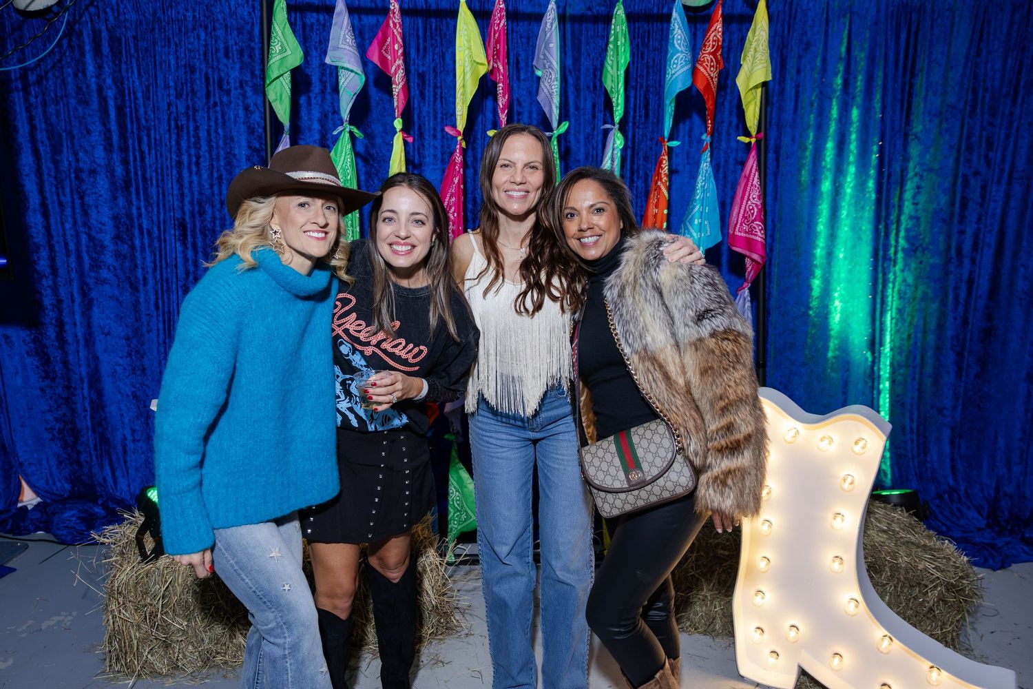 Four women posing together at a colorful western-themed party with hay bales and decorations.